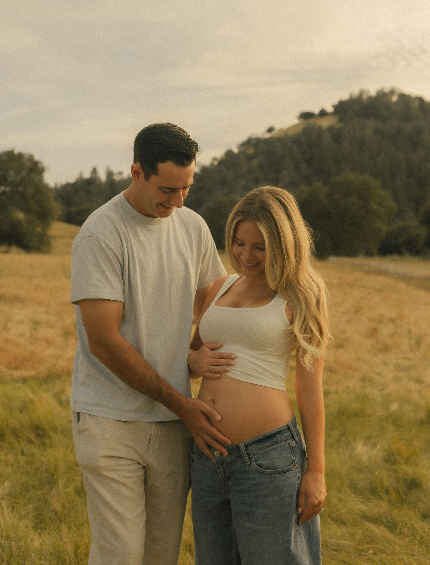 Couple posing in golden field for maternity photo shoot, woman in white crop top showing baby bump.