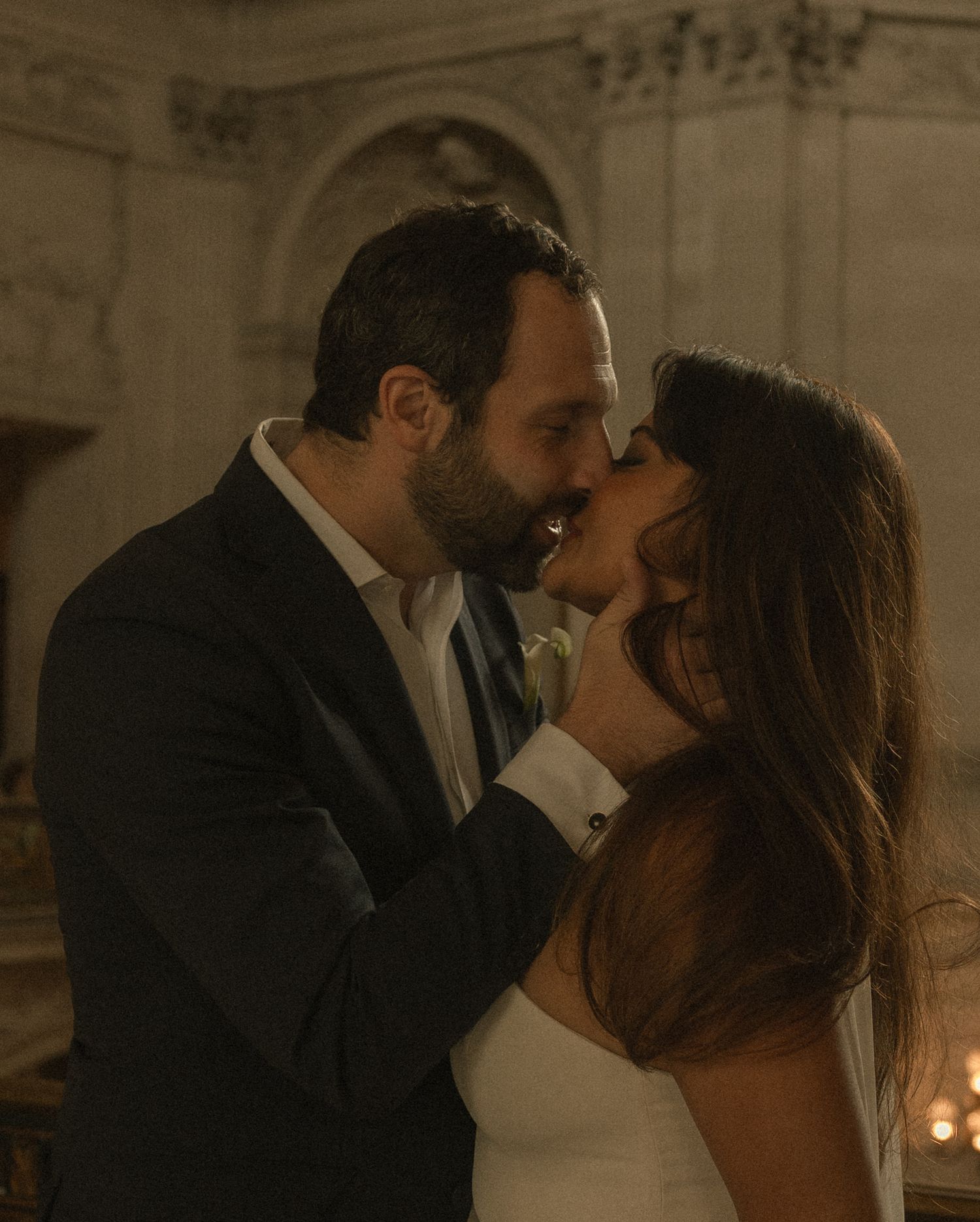 A couple shares a romantic almost-kiss moment in elegant wedding attire inside a grand ornate venue.