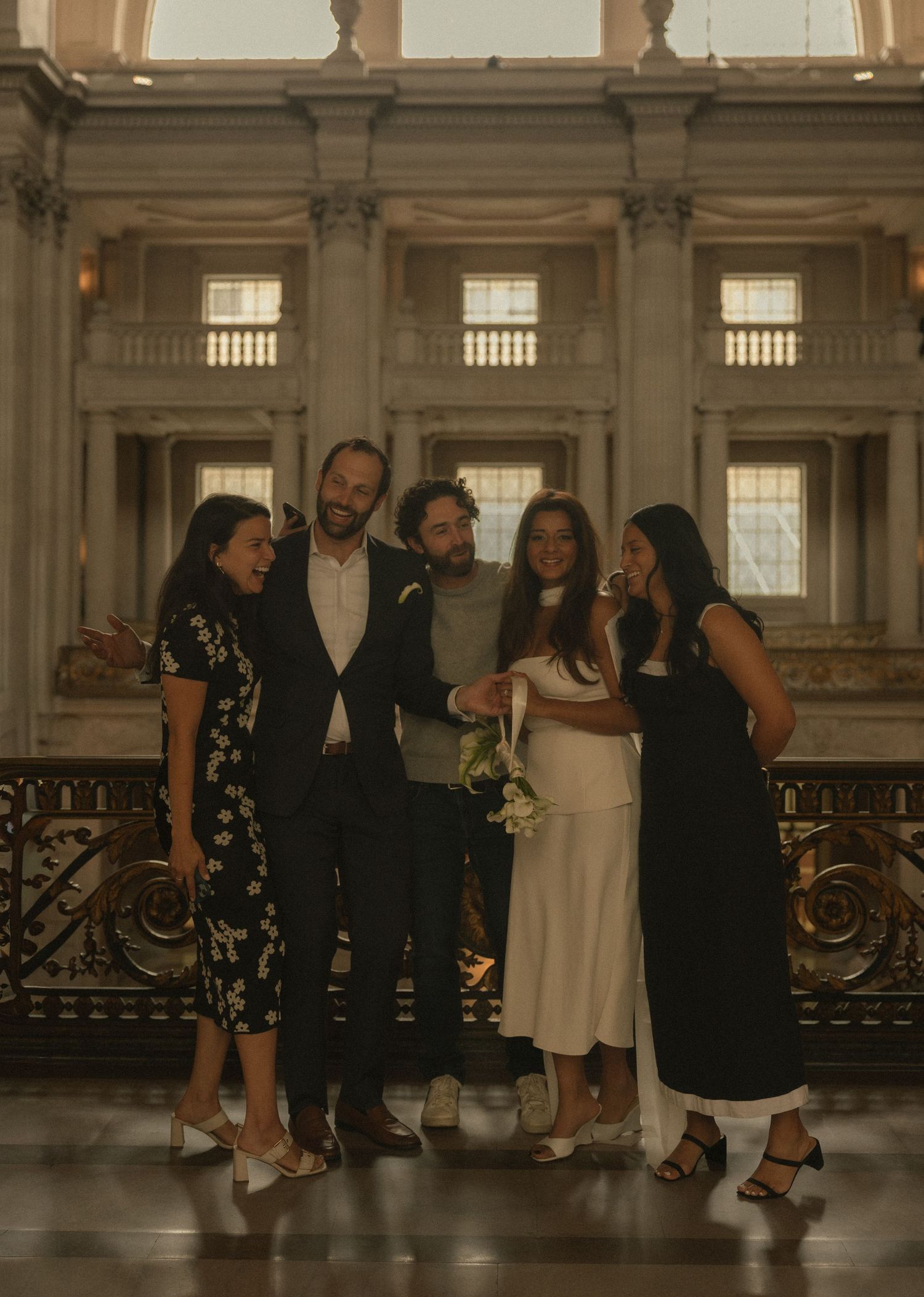 A wedding party of five poses together inside an elegant building with ornate architecture and golden railings.