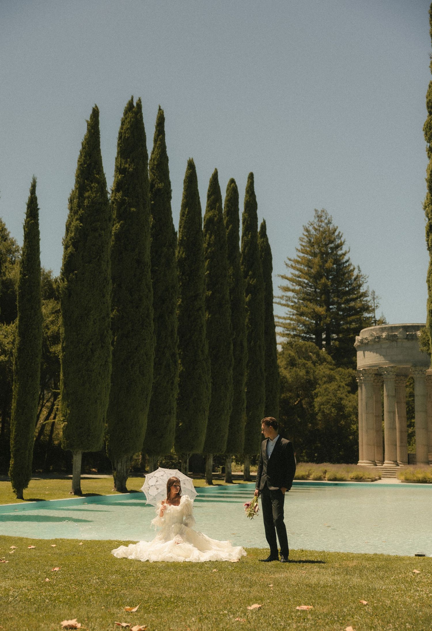 Bride in white gown with parasol sits on grass beside groom near cypress trees and classical columns at outdoor wedding.