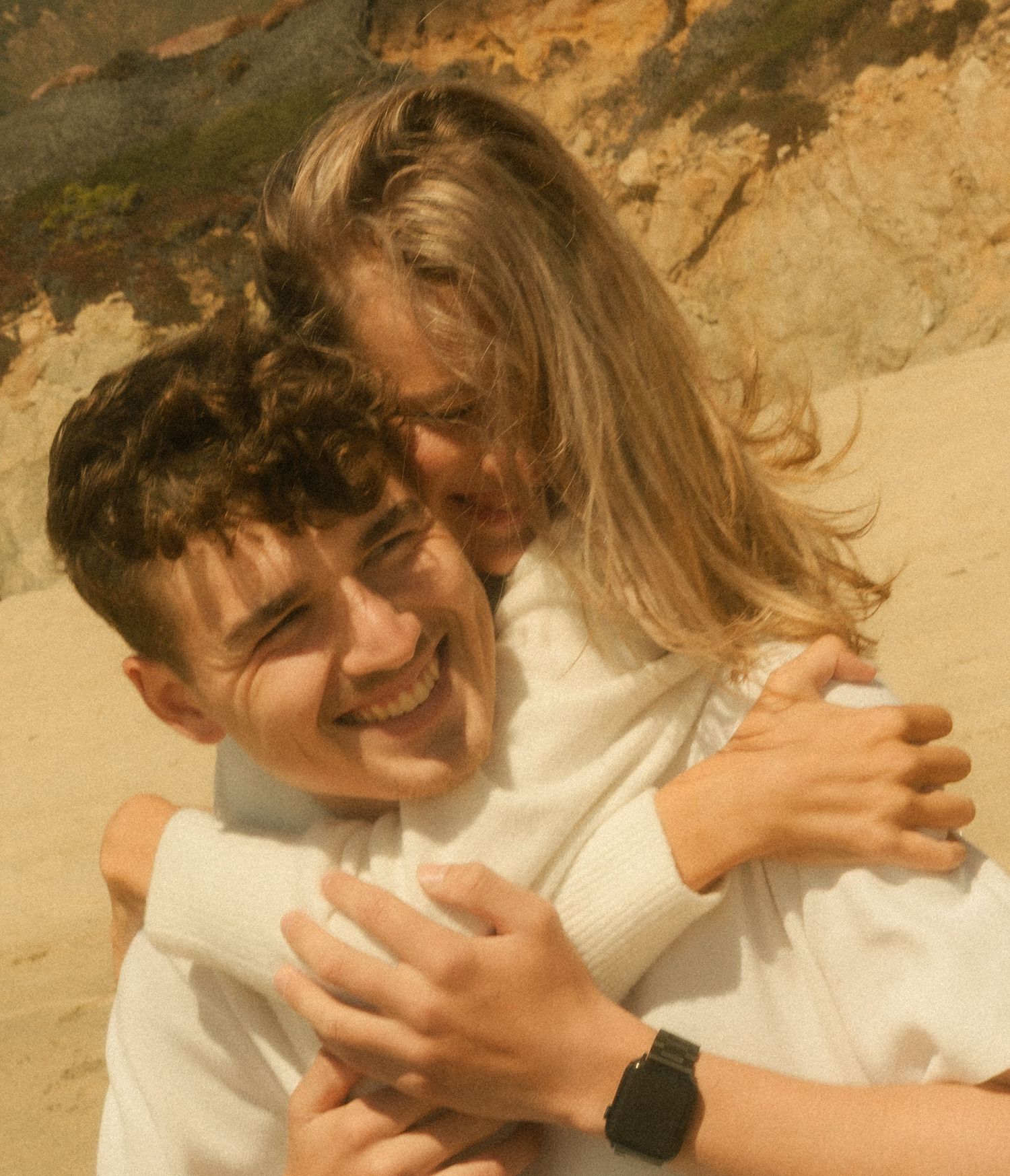 Two young people hugging and smiling on a sandy beach, both wearing white sweaters on a sunny day.