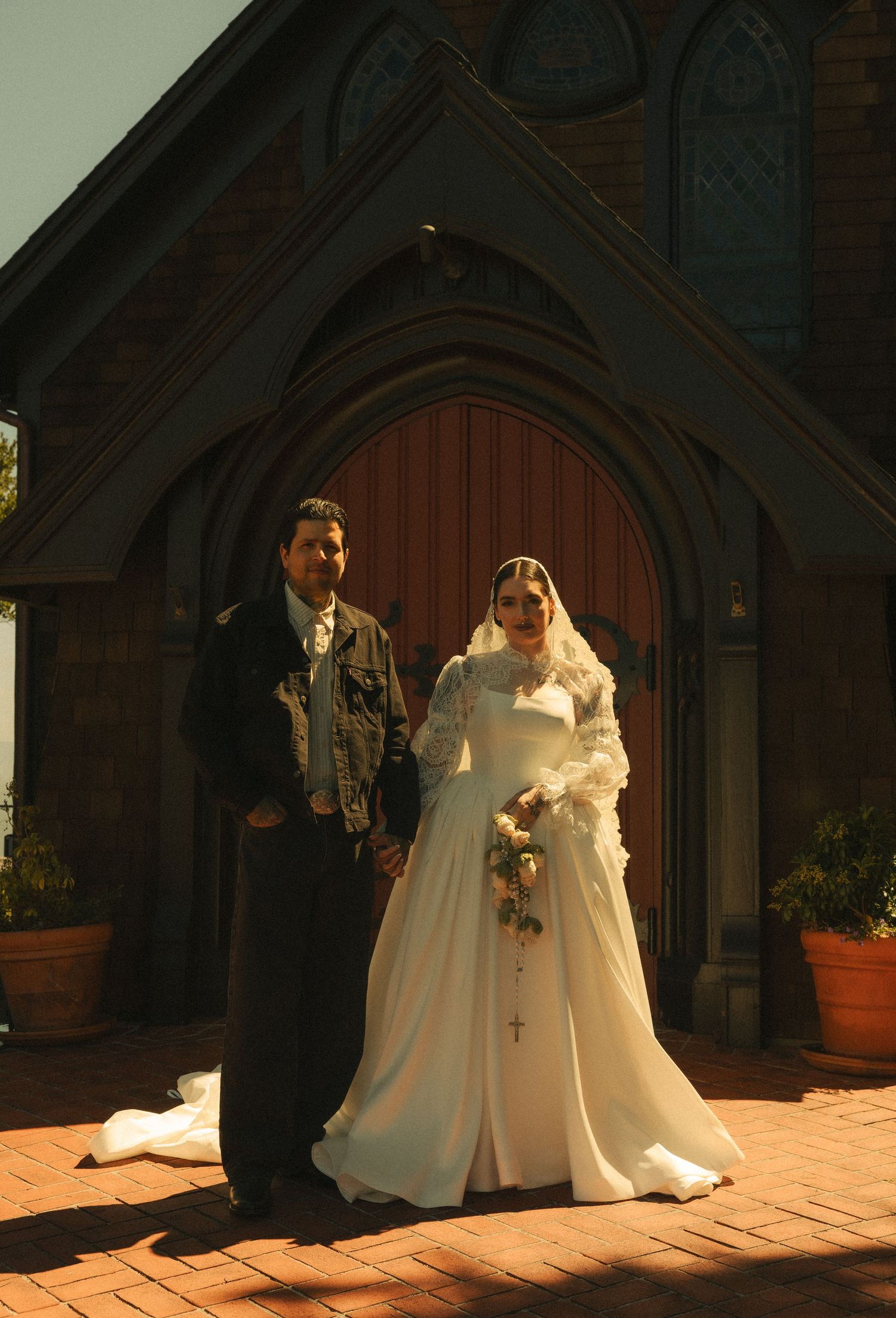 Bride and groom posing in front of a brick church with arched wooden doors on their wedding day.
