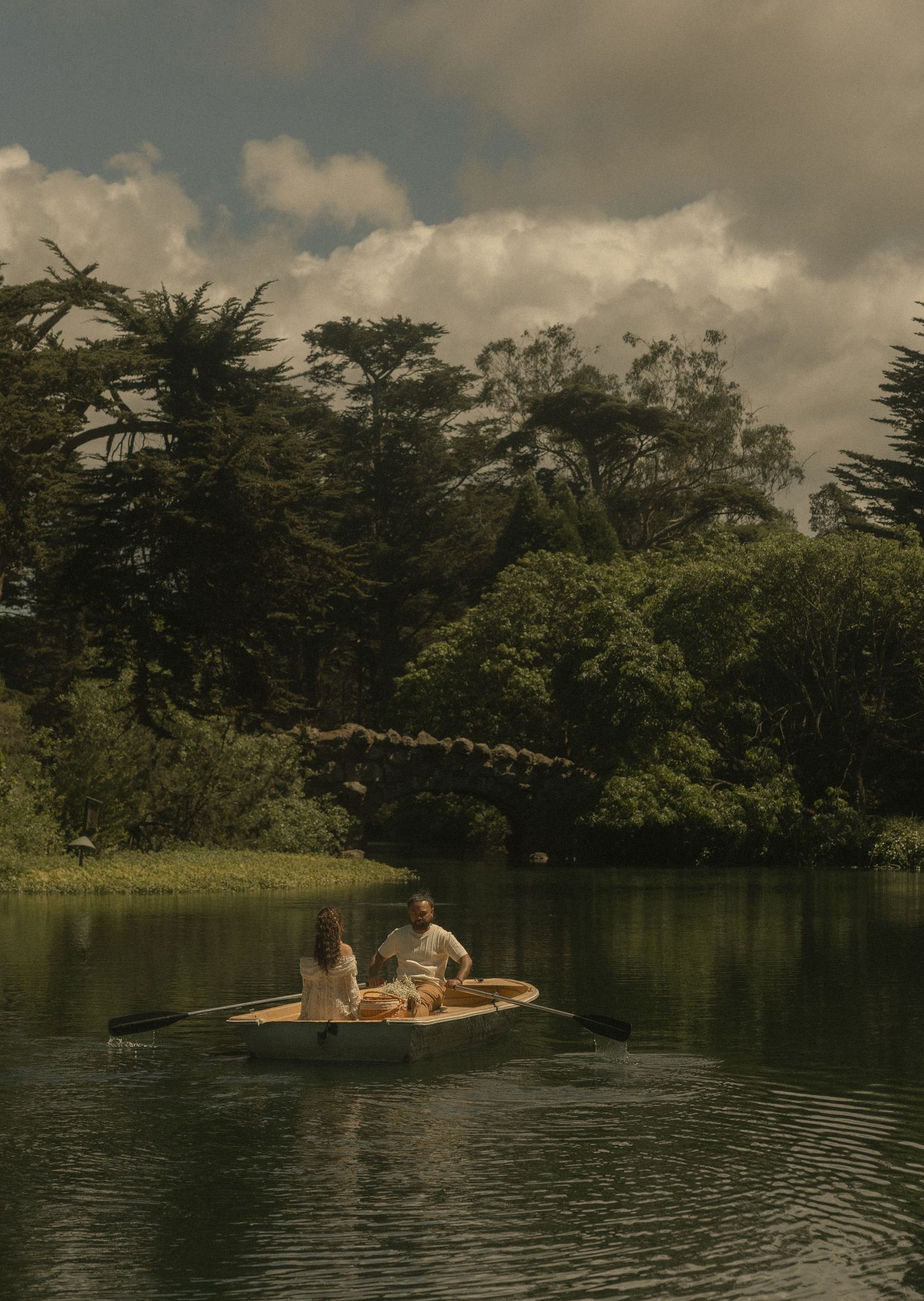 Couple rowing a wooden boat on a serene lake surrounded by lush green trees and a stone bridge.