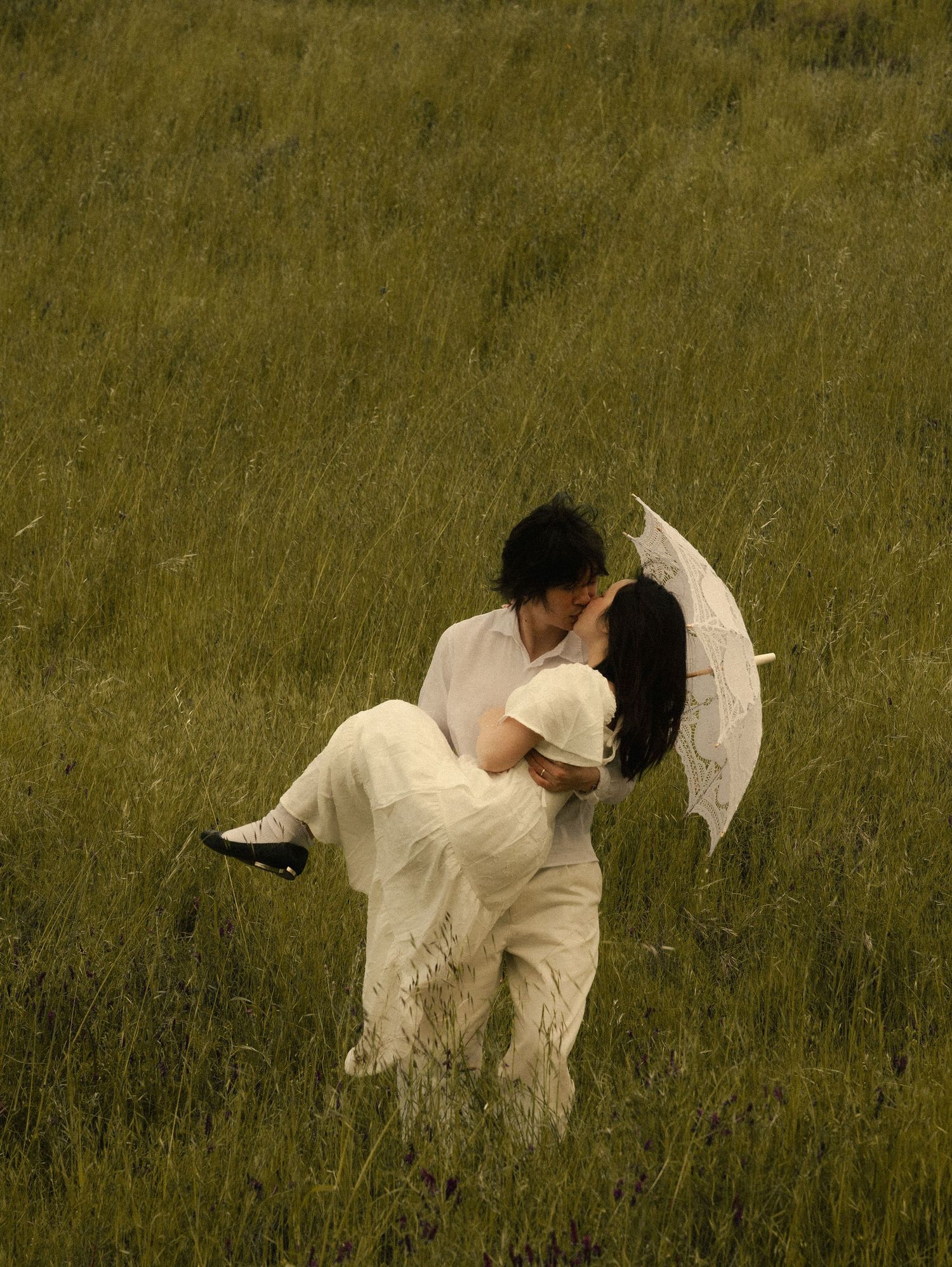 A couple in white clothing share a romantic kiss in a green meadow, with the man lifting the woman holding a lace umbrella.