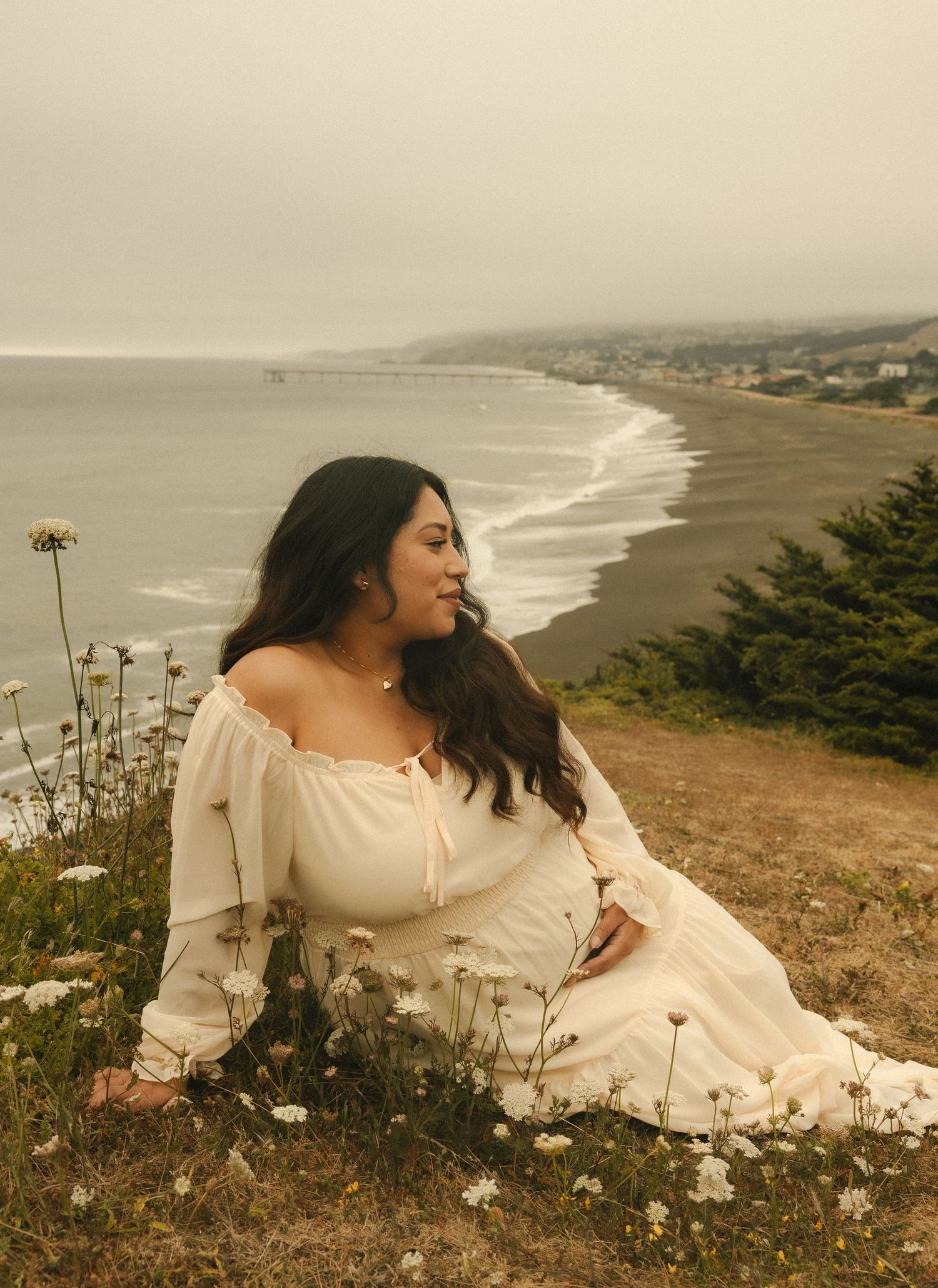 Woman in white off-shoulder dress sits on coastal hillside with wildflowers, overlooking a misty beach shoreline.