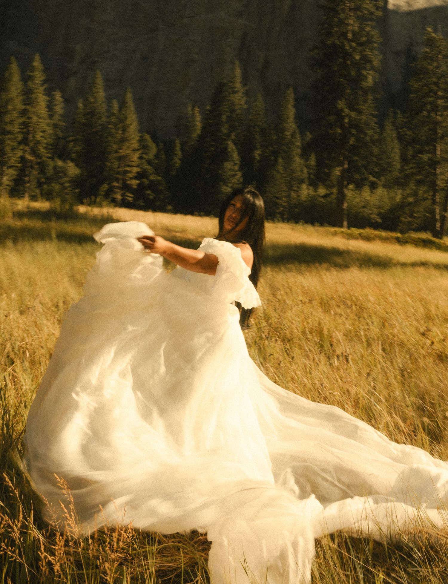 A woman in a flowing white off-shoulder gown twirls in a golden meadow with pine trees and mountains behind her.