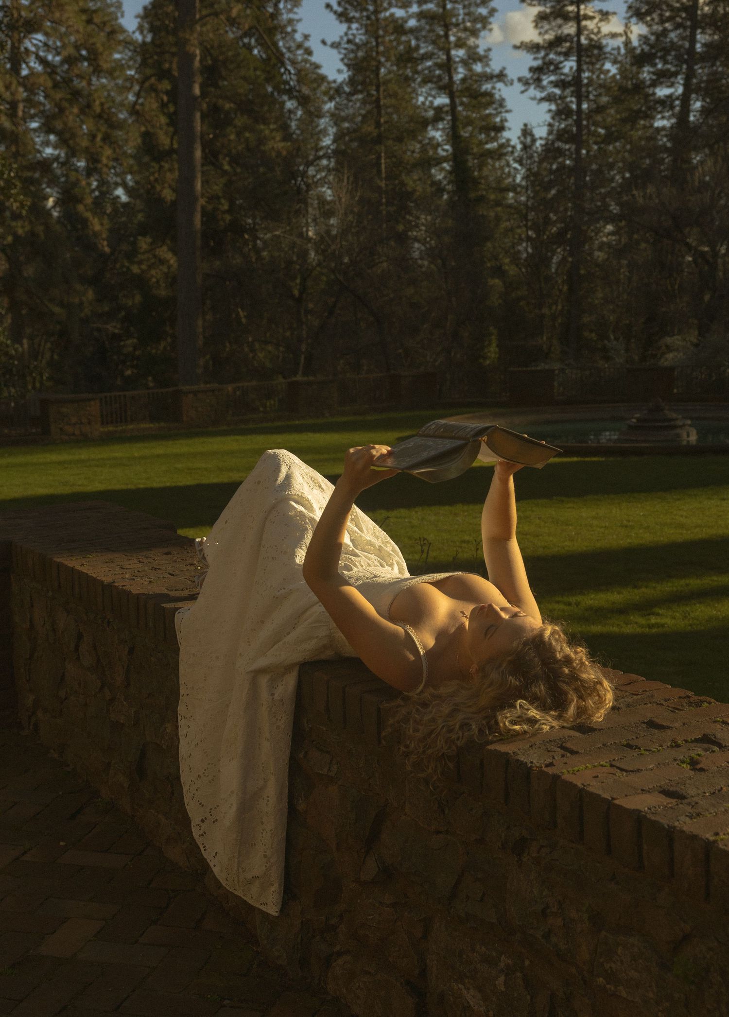 Figure in white dress reclining on stone wall at sunset with forest backdrop.
