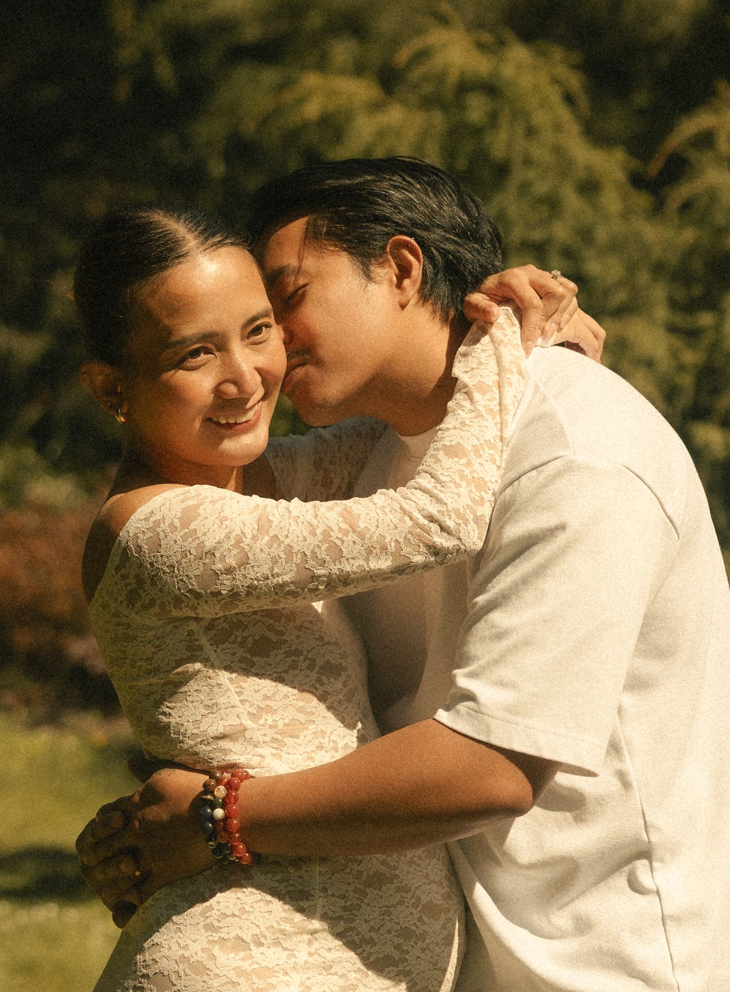 A couple embraces outdoors, woman in lace dress smiling as man kisses her cheek in a sunlit garden.