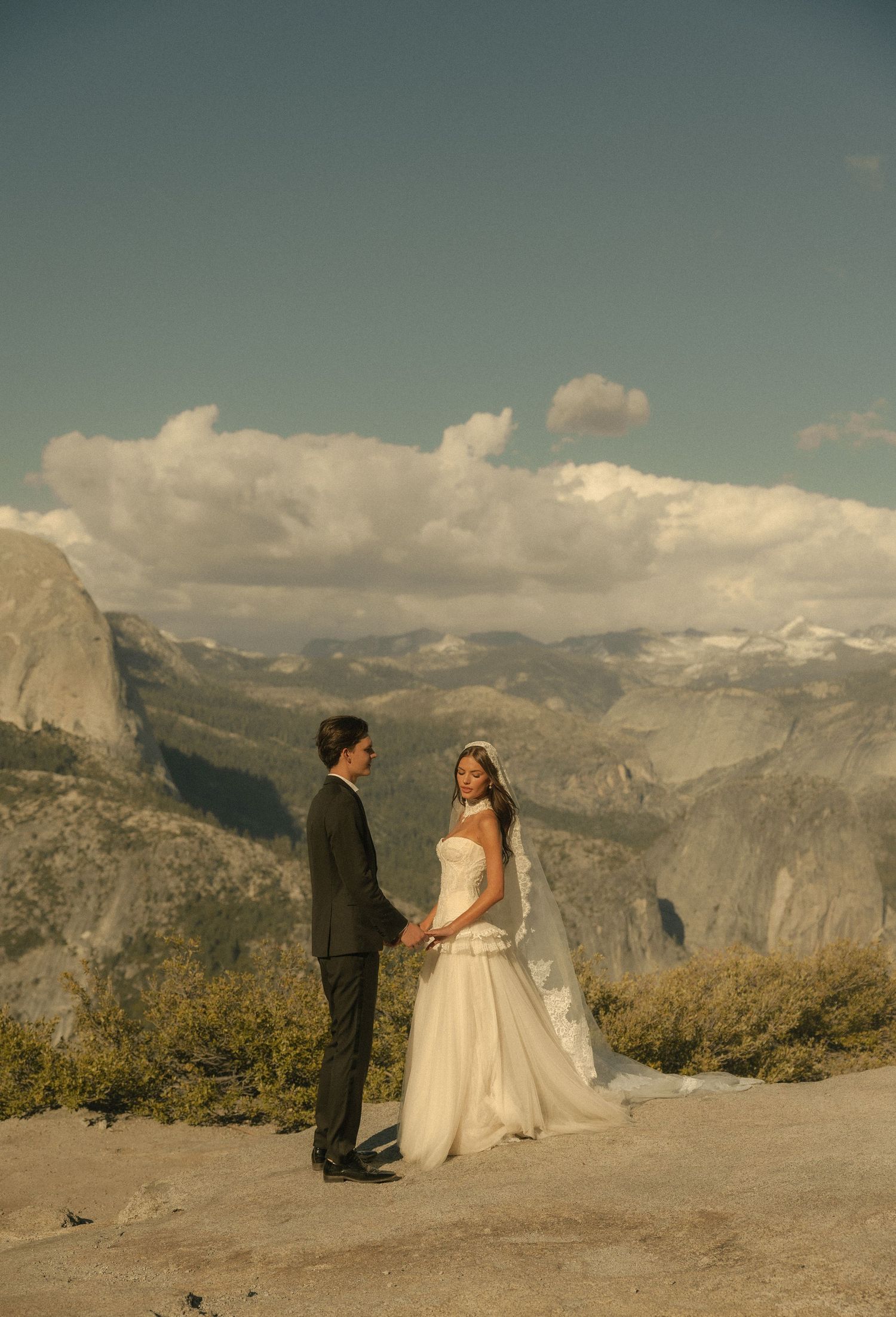 Bride and groom hold hands at Glacier Point, Yosemite, with Half Dome and mountain views behind them.