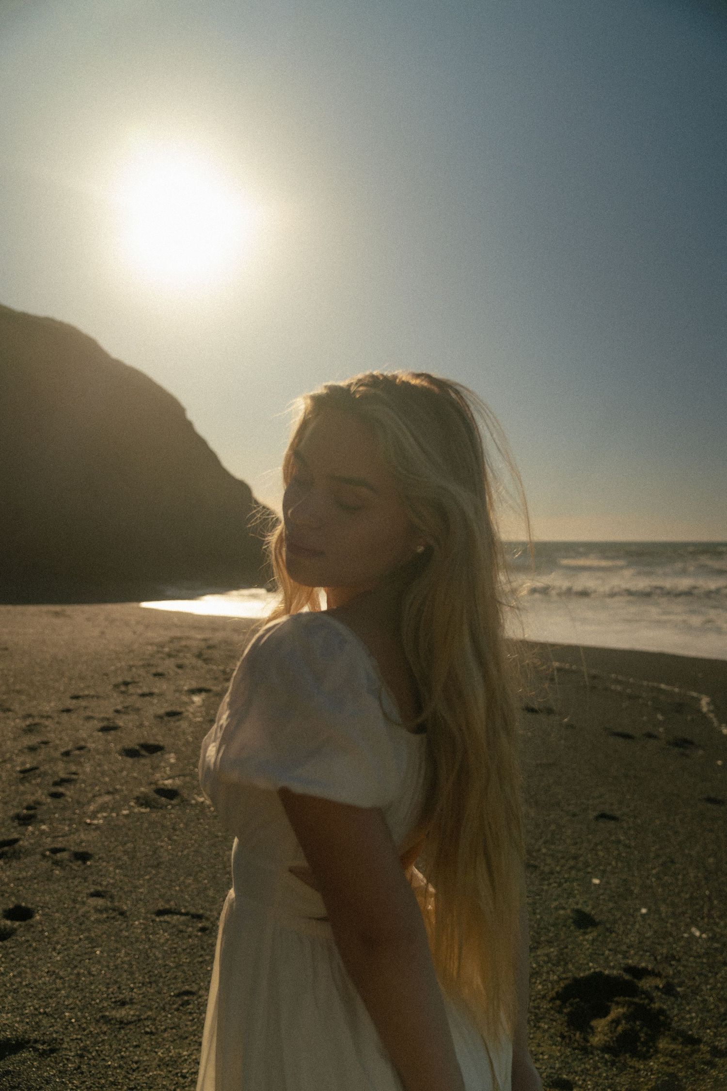 Blonde woman in white dress standing on a sandy beach at golden hour with ocean waves and rocky cliffs.