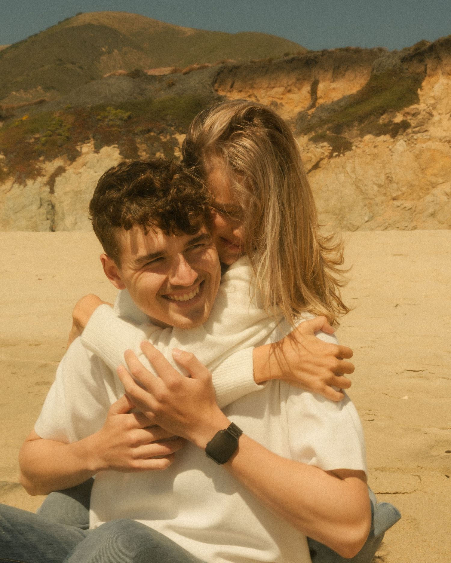 Two young people hugging and smiling on a sandy beach with rocky cliffs in the background on a sunny day.