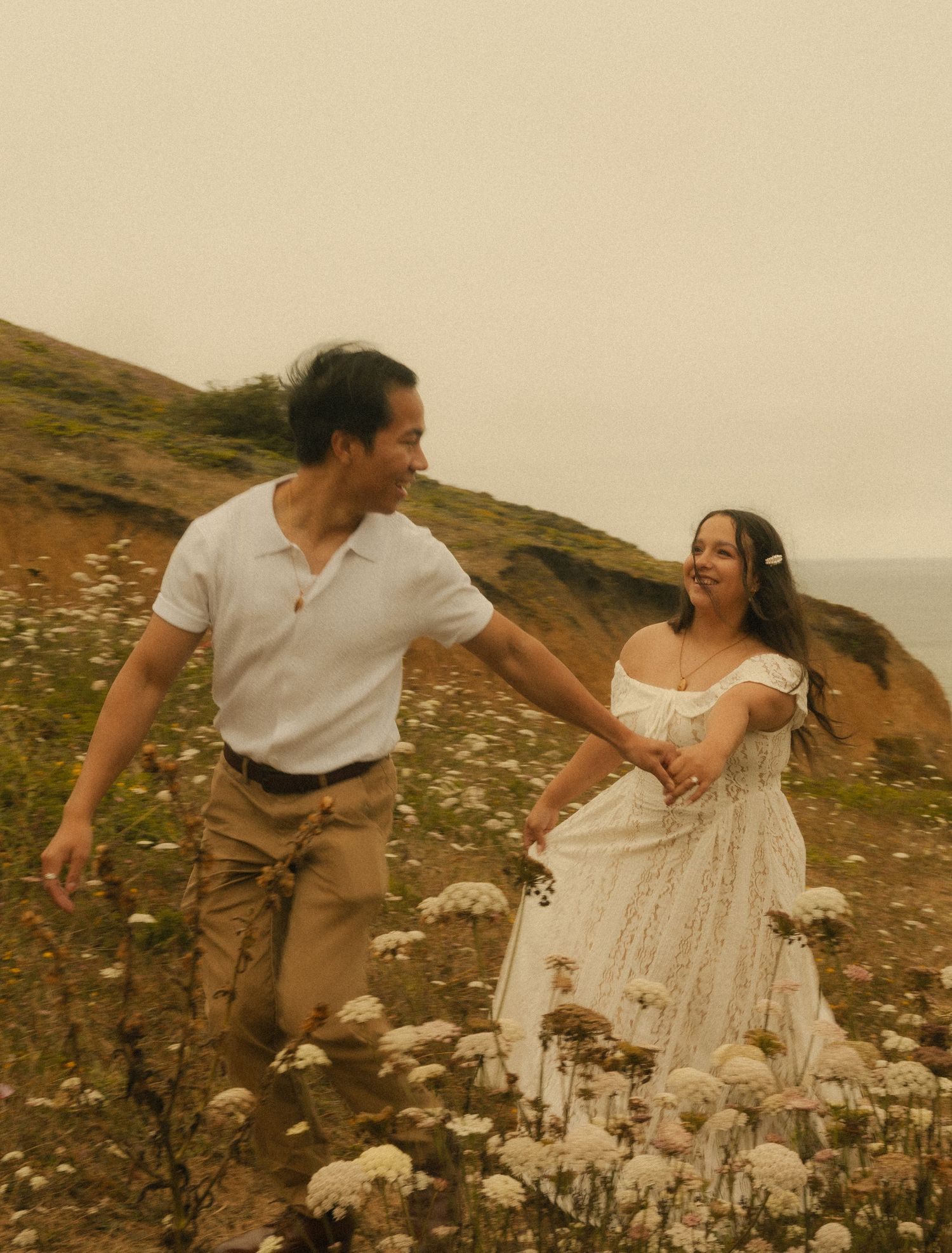 Couple holding hands walking through wildflowers on a coastal cliffside, woman in white dress, man in white shirt and khakis.