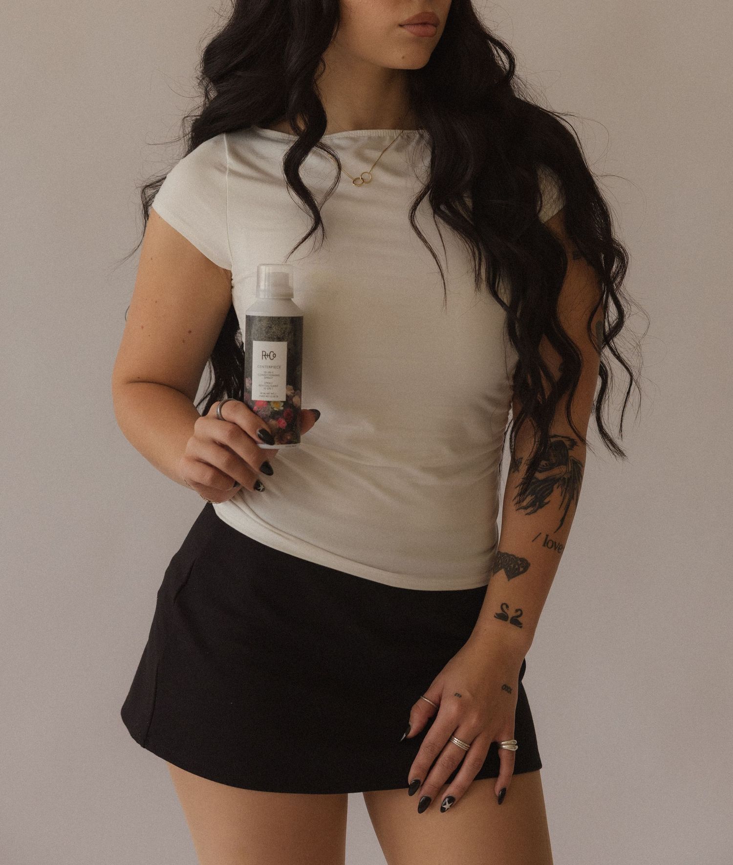 Woman with long dark hair, tattoos, wearing white ribbed tee and black mini skirt, holding a hair product bottle.