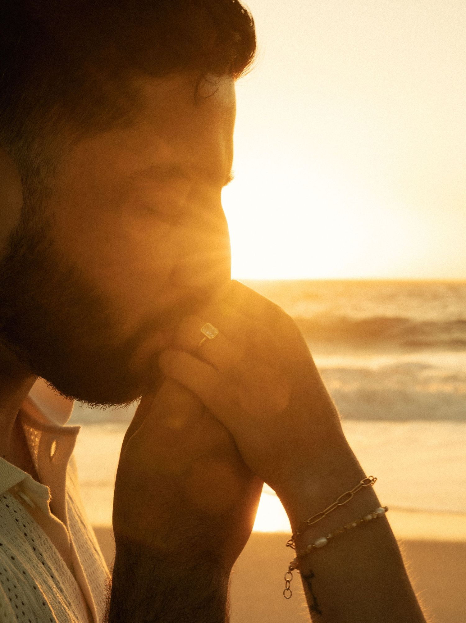 Bearded man at golden hour sunset on beach, warm sunlight flare, wearing bracelet, contemplative mood.