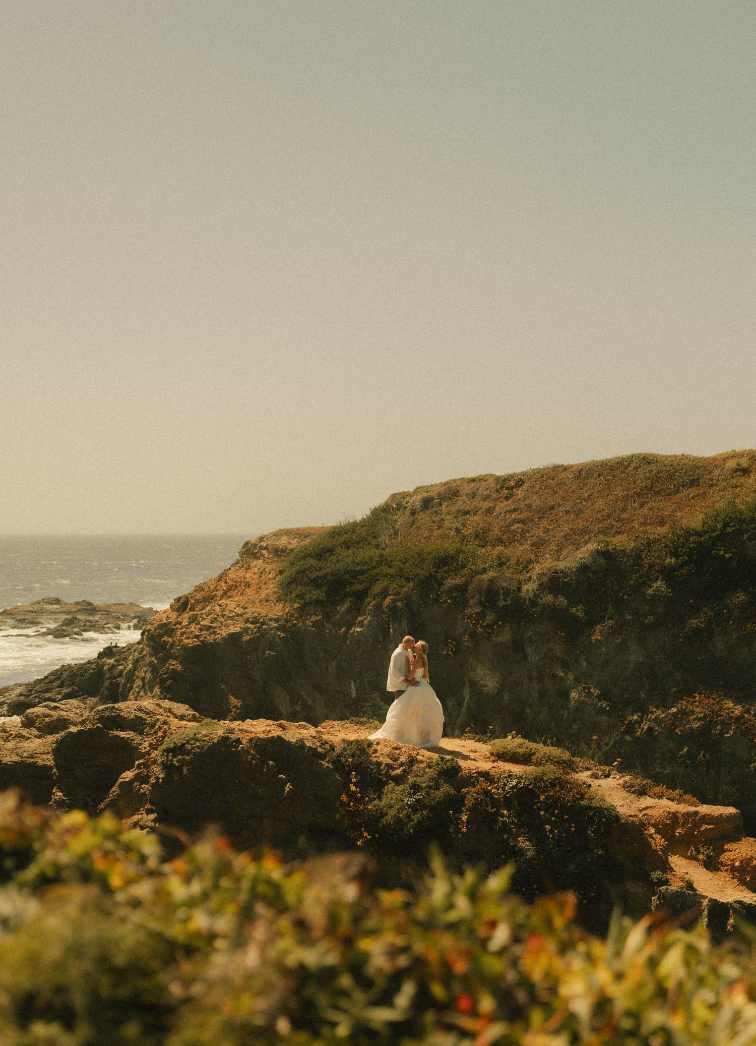 A bride and groom embrace on rocky coastal cliffs overlooking the ocean on their wedding day.