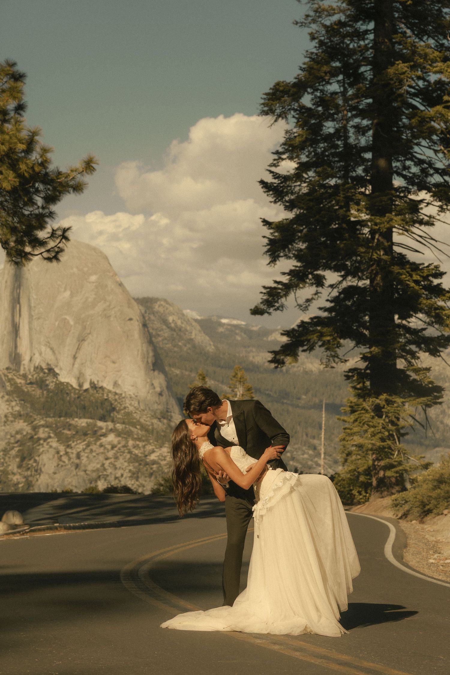 Couple sharing a romantic dip kiss on a winding mountain road with tall pines and rocky peaks behind them.
