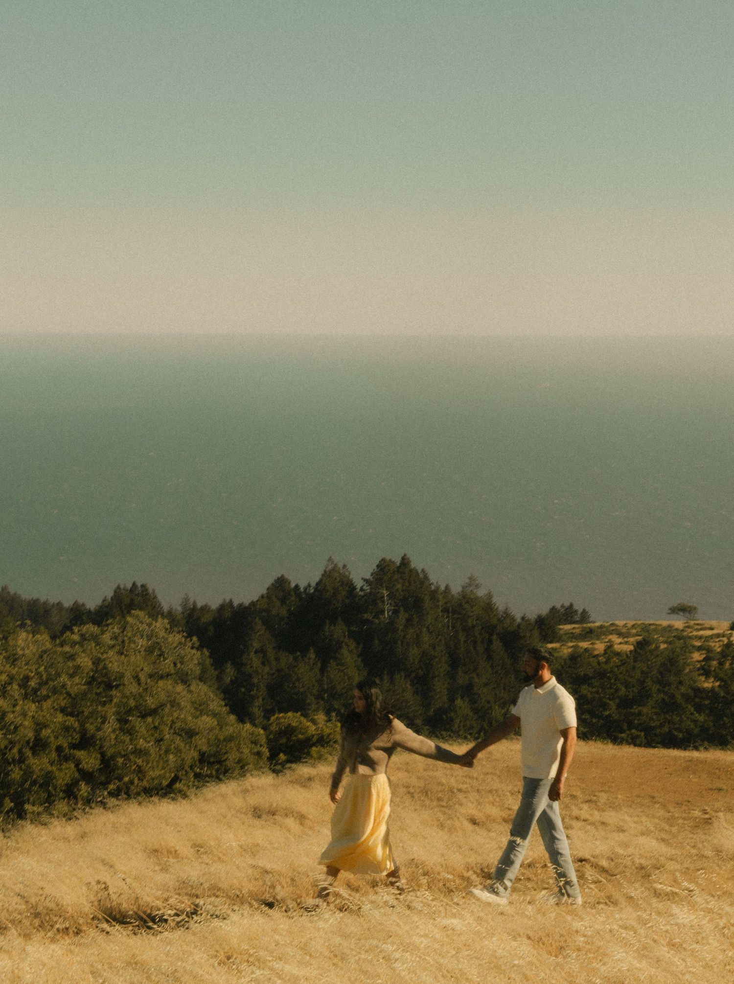 Couple holding hands walking on golden hillside overlooking ocean and forest on a clear sunny day.