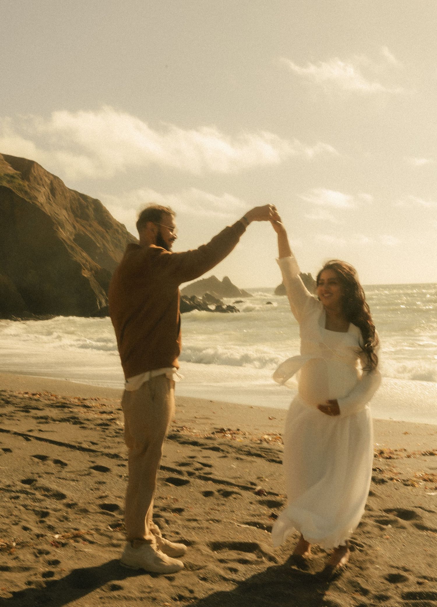 Couple dancing on beach at sunset, woman in white dress with baby bump, man in brown sweater.