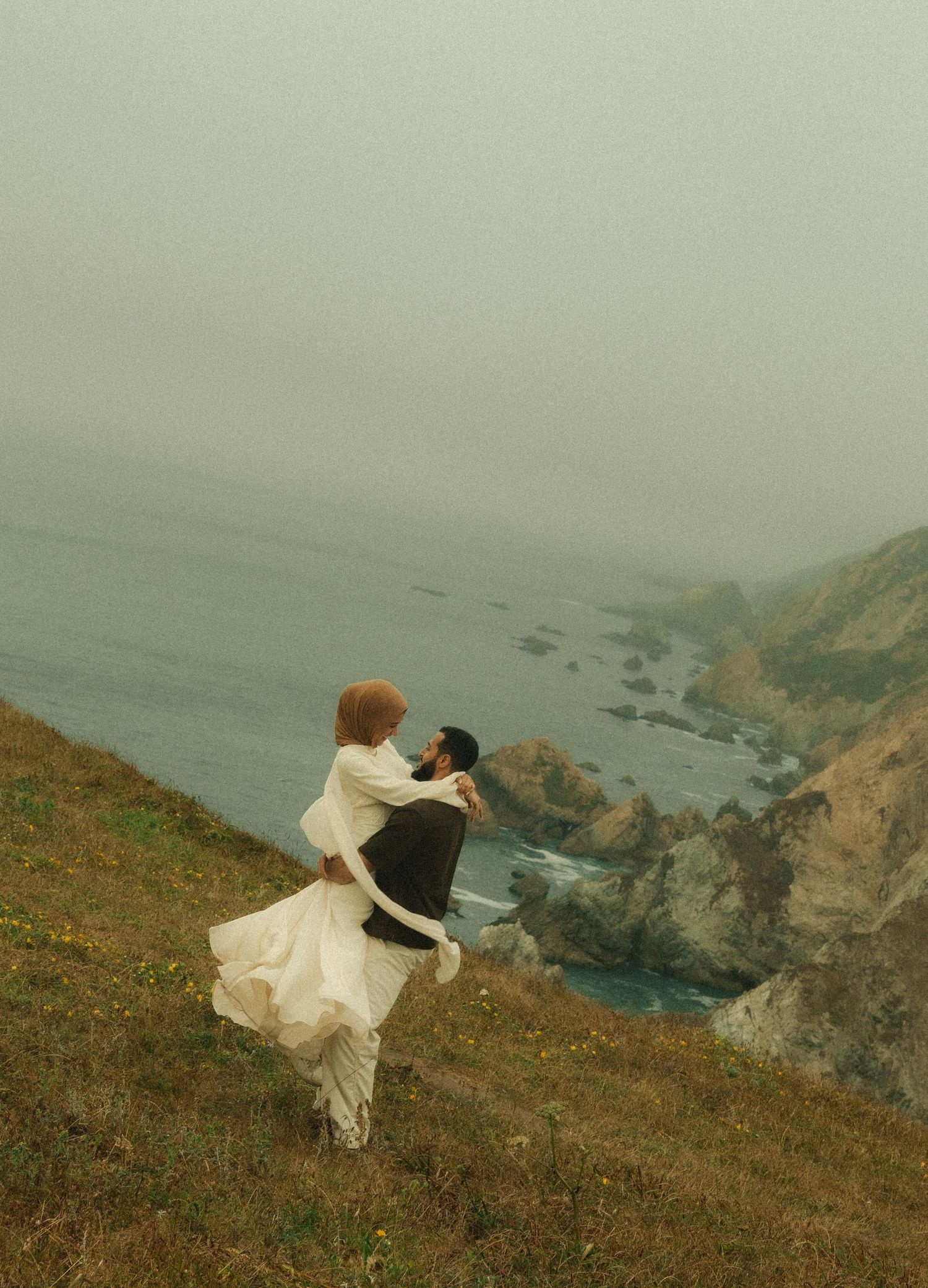 Couple dancing on a misty coastal hillside cliff overlooking the ocean, wearing white and black attire.