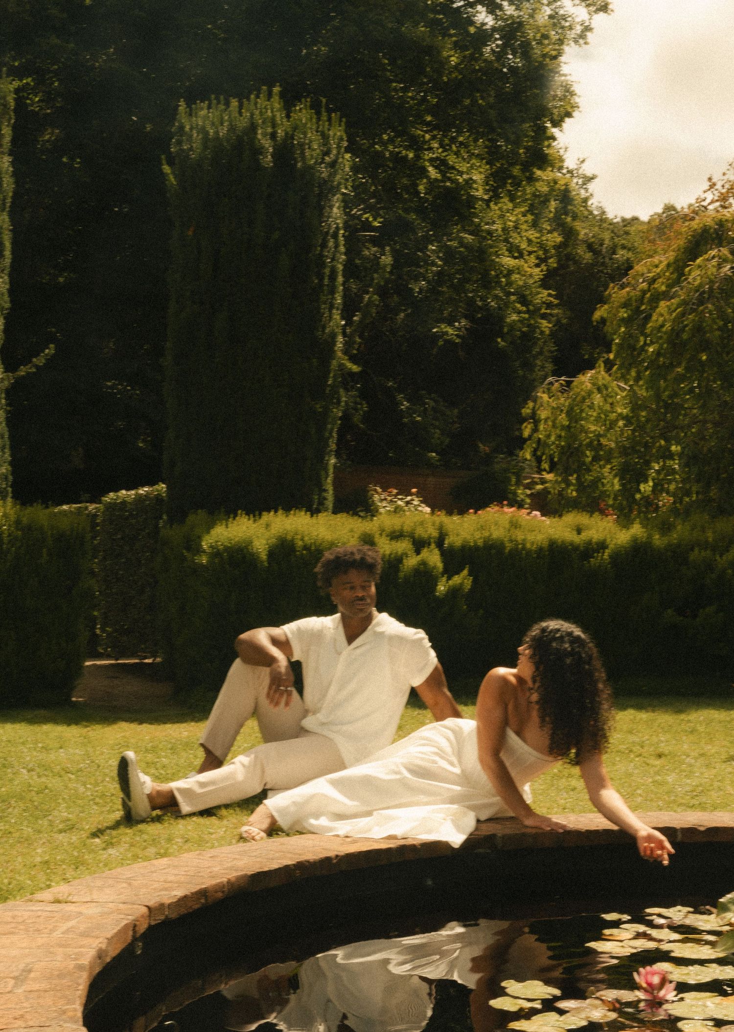 Couple in white outfits relaxing by a lily pond in a lush green garden on a sunny day.