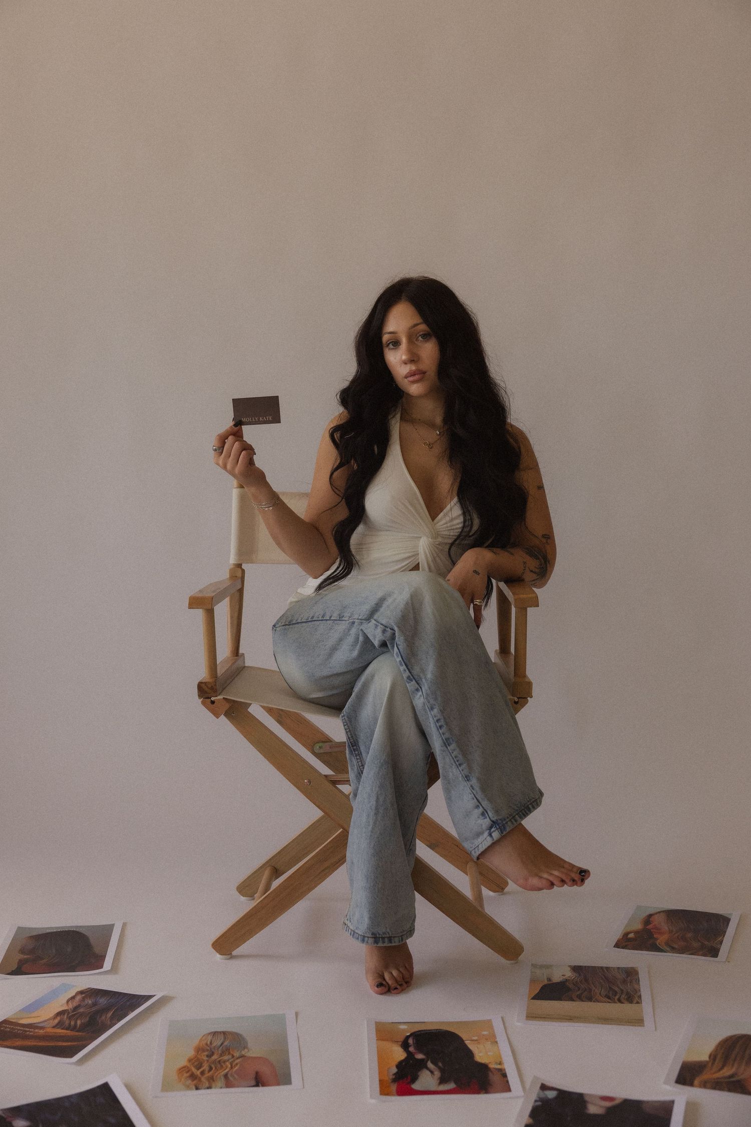 Woman with long dark hair sits in a director's chair holding a photo, surrounded by scattered polaroids on white background.