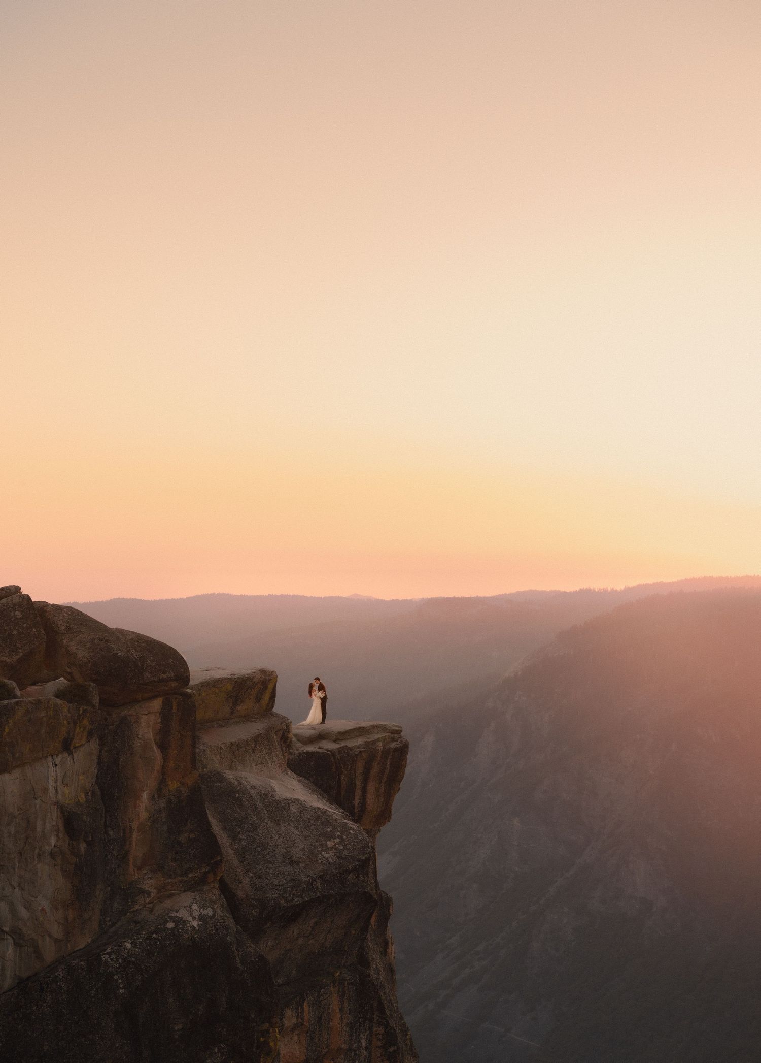 Couple in wedding attire standing on a rocky cliff edge at sunset with mountain valley views in Yosemite.