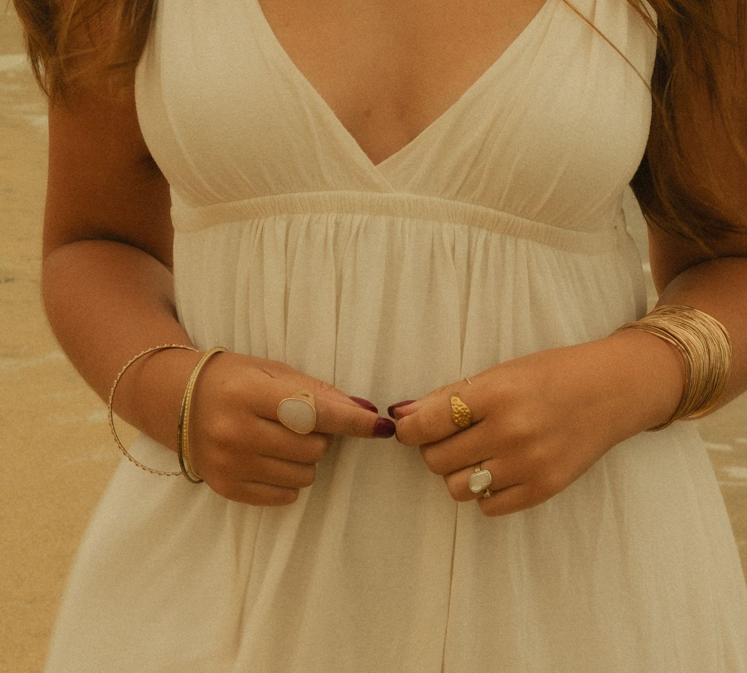 Woman in white V-neck dress wearing gold bracelets and rings with dark red nail polish, hands clasped at waist.