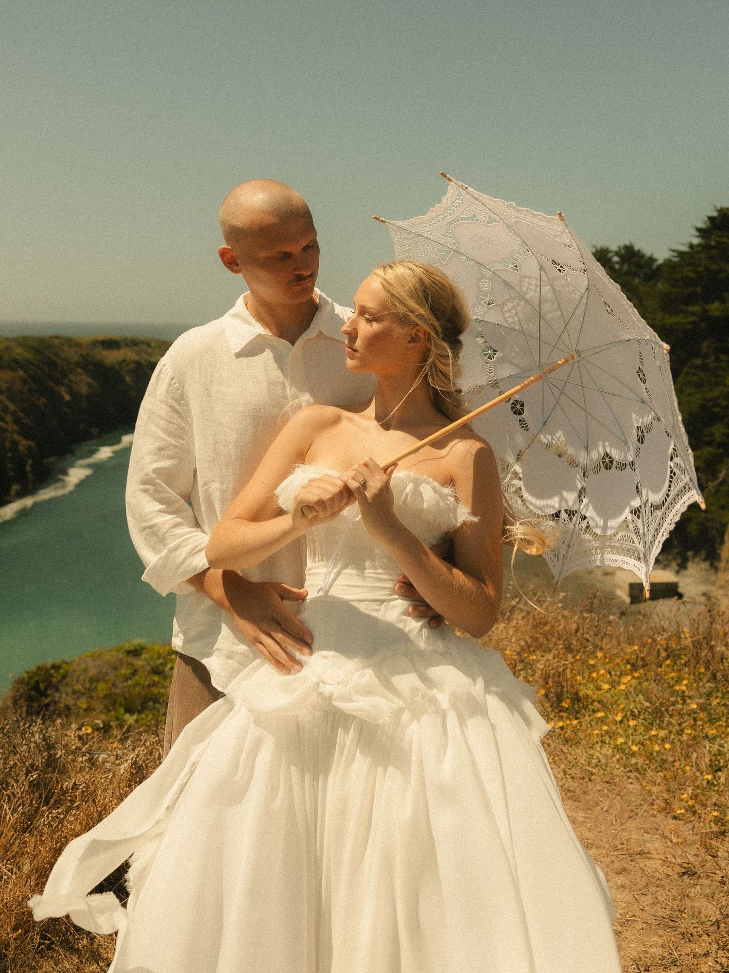 Bride in white gown holding lace parasol with groom on coastal cliffside, ocean view wedding portrait.