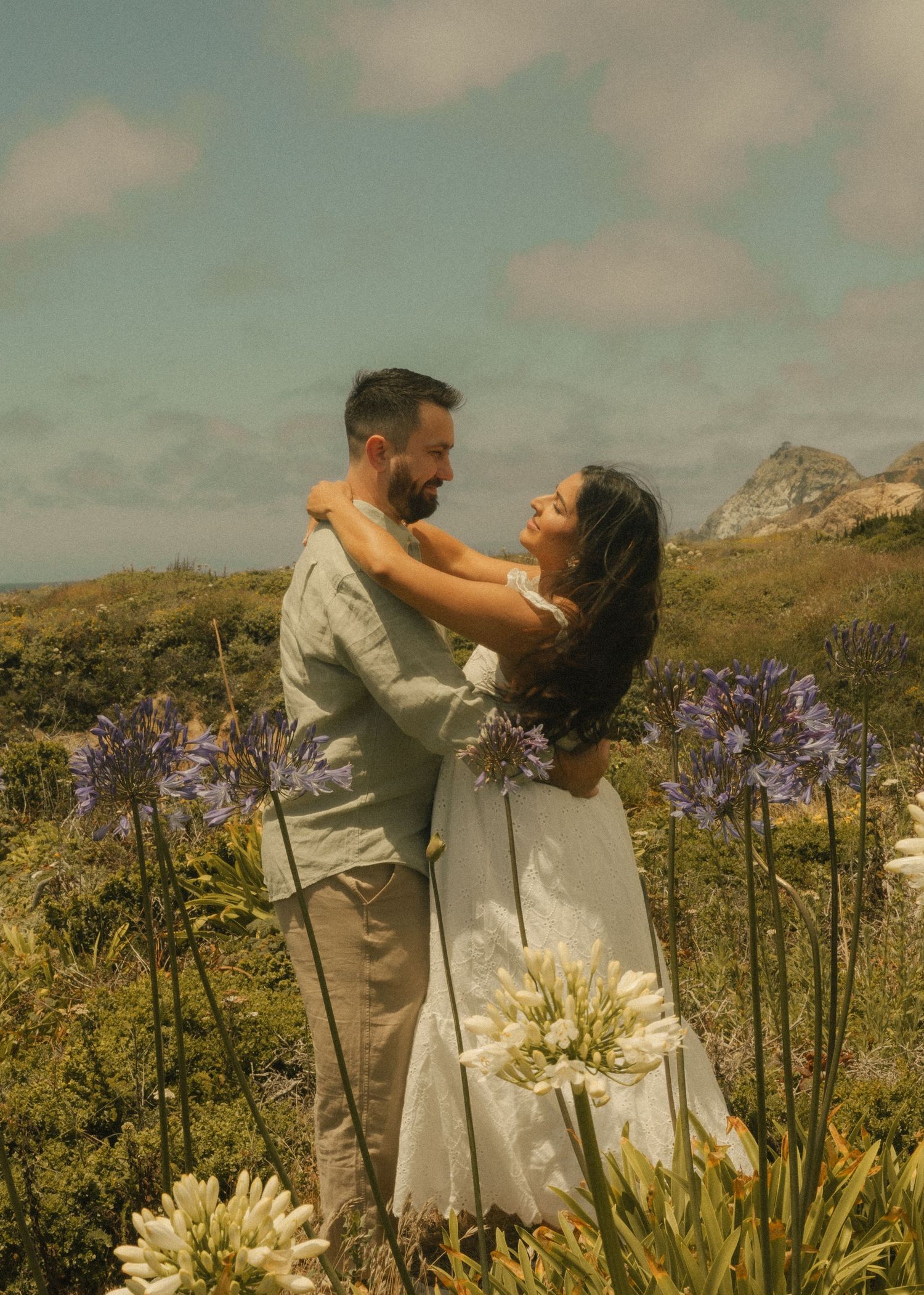 Couple embracing among purple agapanthus flowers with mountain backdrop on a sunny day.