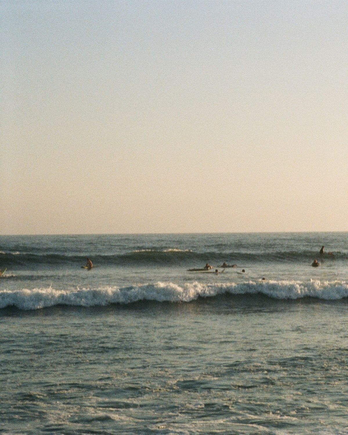 Ocean waves rolling in with surfers visible in distance during sunset.