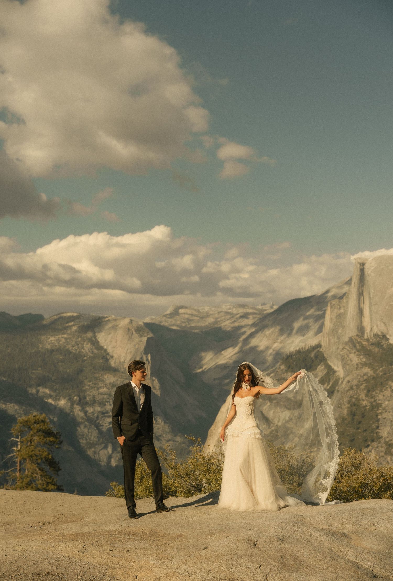 Bride and groom stand on rocky overlook at Yosemite with Half Dome backdrop, veil flowing in wind.