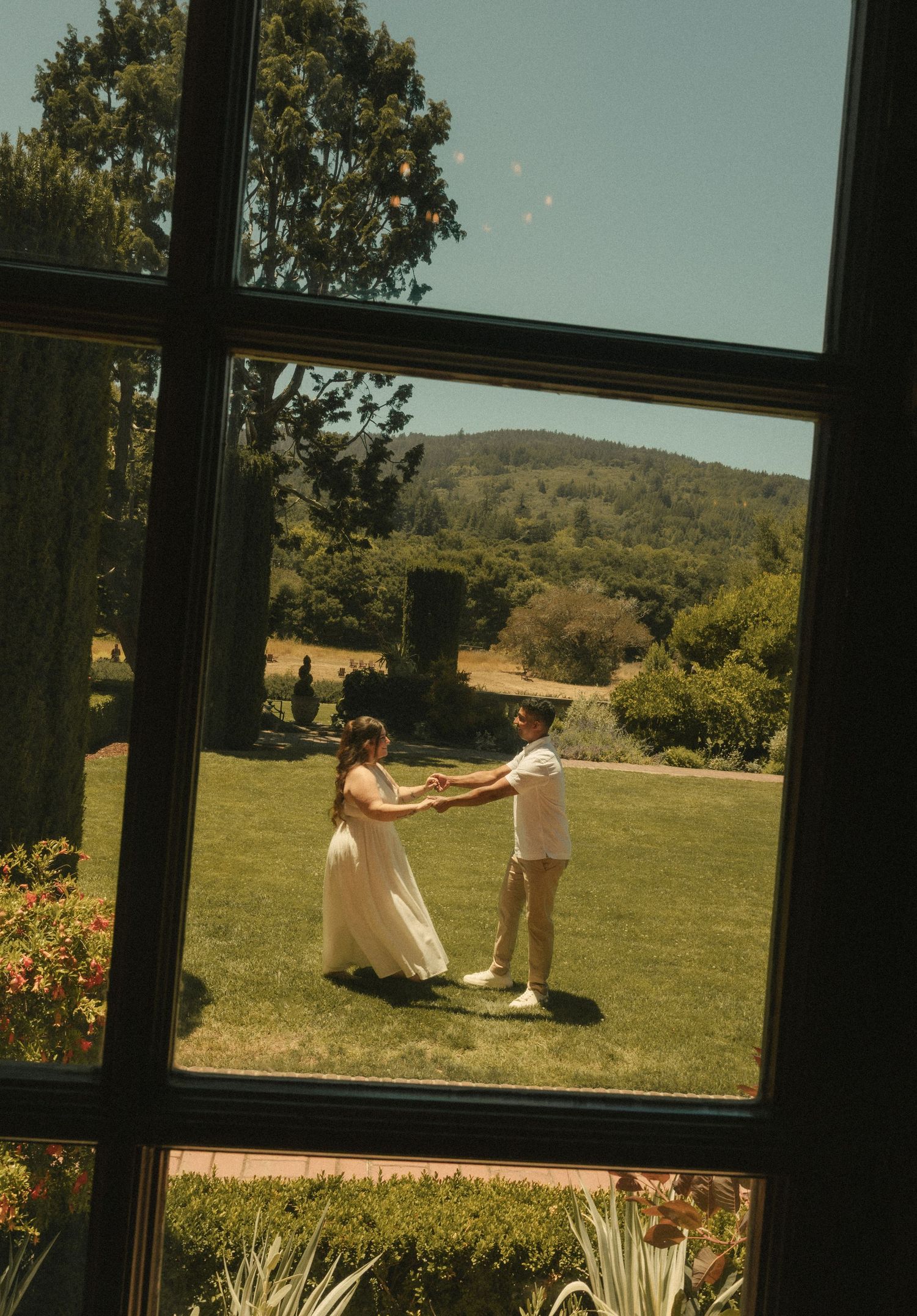 Couple dancing on lush green lawn viewed through window, with rolling hills and trees in background on sunny day.