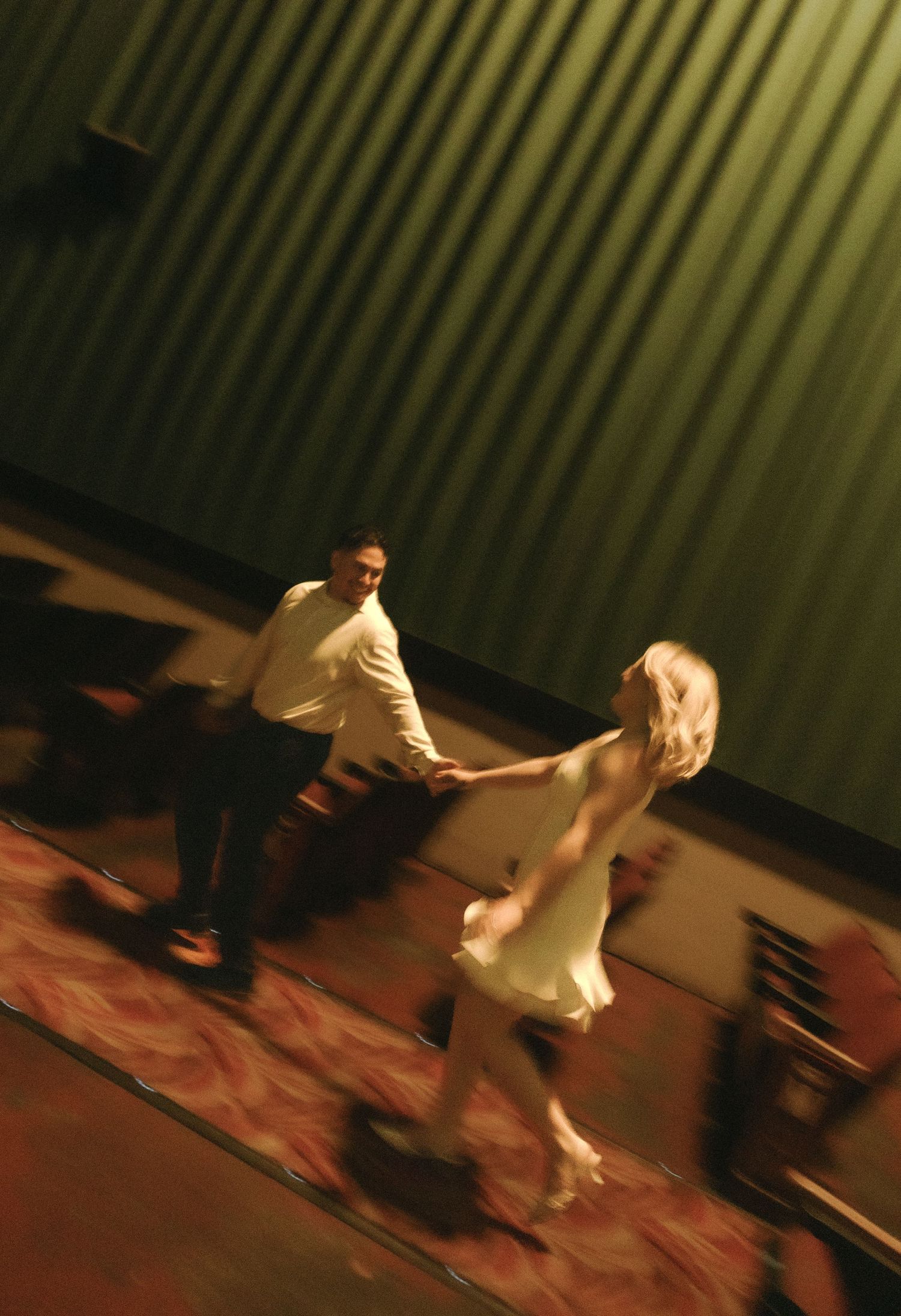A couple dances energetically on a patterned floor, the woman in a white dress, captured with motion blur.