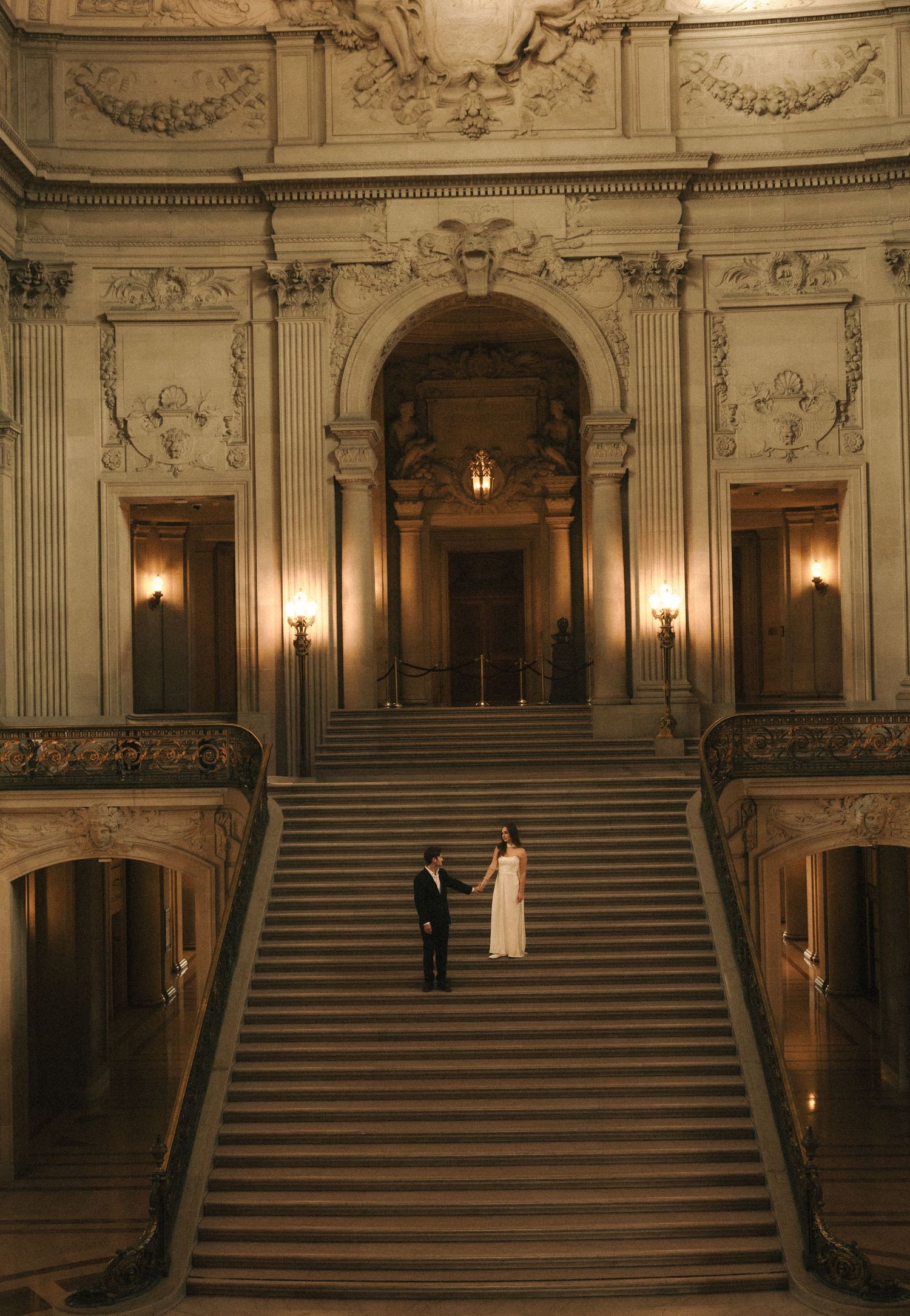 Elegant wedding couple standing on grand staircase inside San Francisco City Hall with ornate Beaux-Arts architecture.