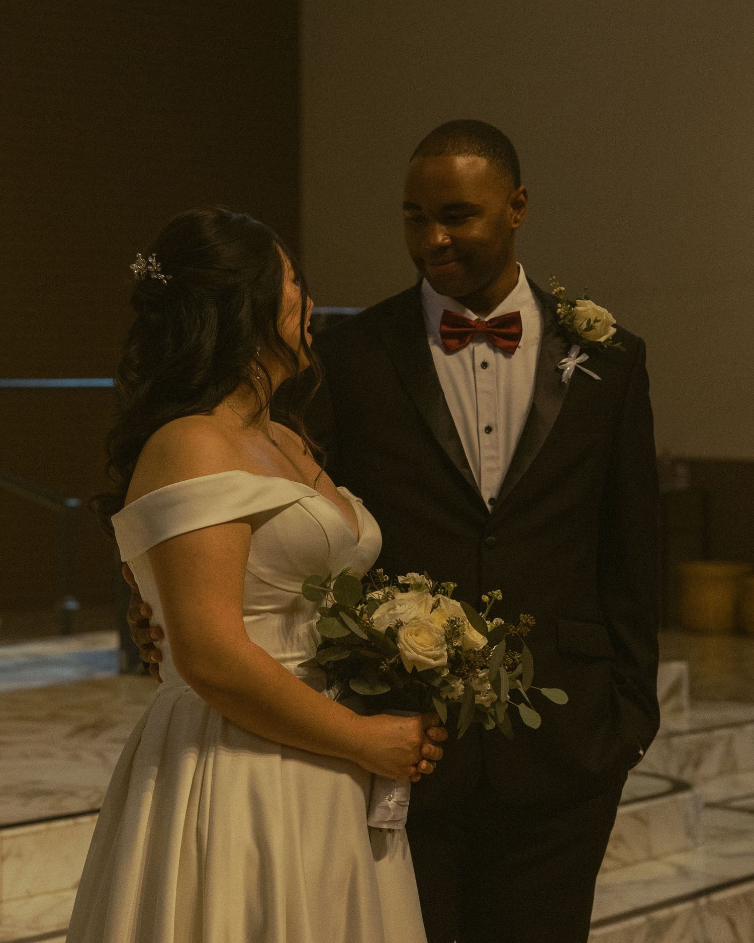 A couple shares an intimate moment on their wedding day in formal attire against a dark indoor backdrop.