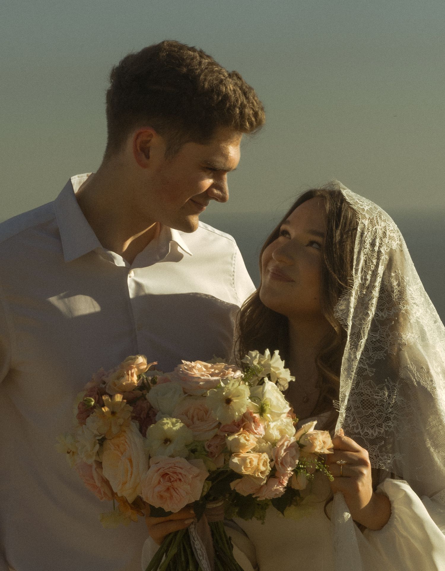 A couple in wedding attire share an intimate moment at sunset while holding a bouquet of pink and white flowers.