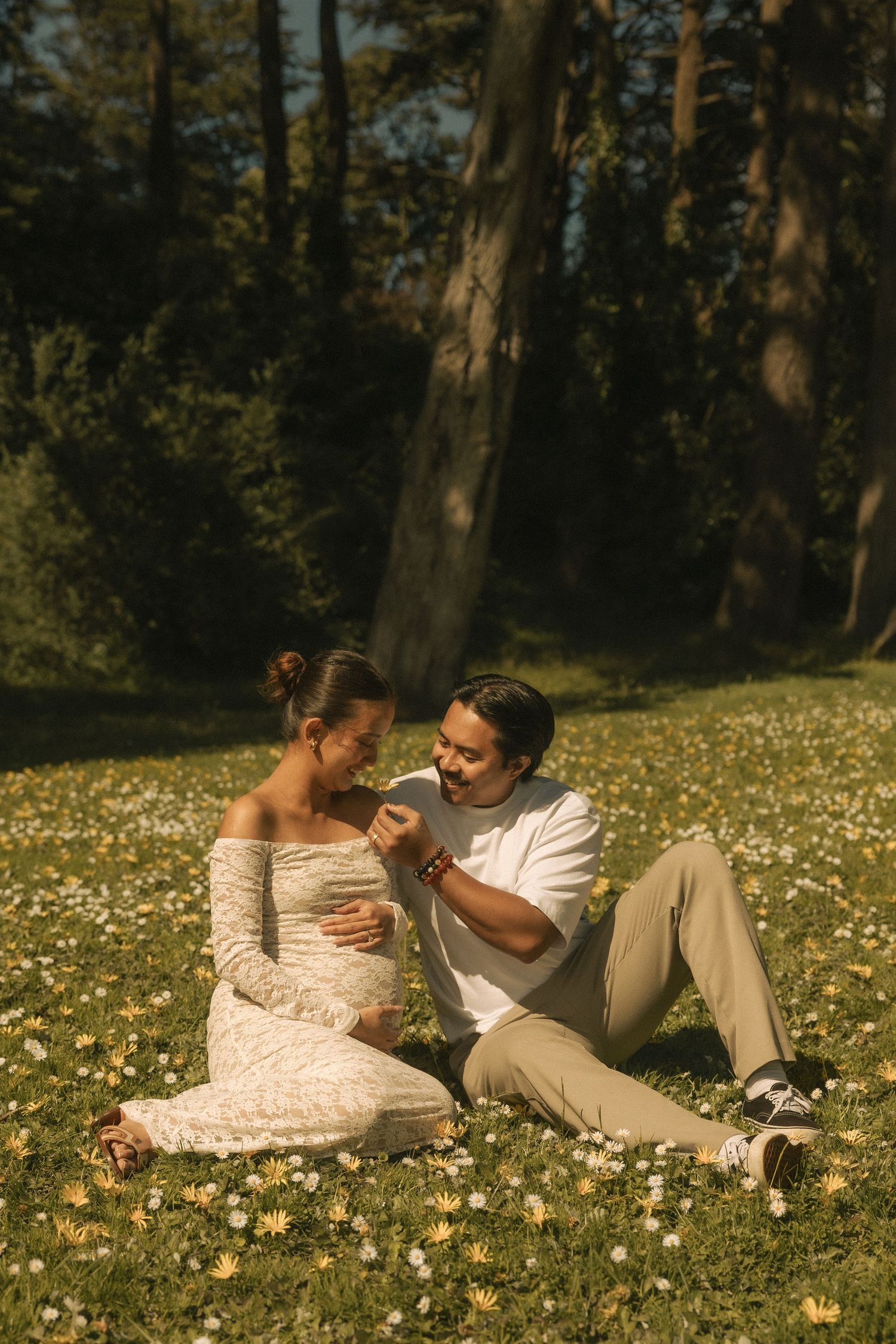 Pregnant couple sitting in a sunny meadow with wildflowers, man tenderly holding woman's hand in a lush park.