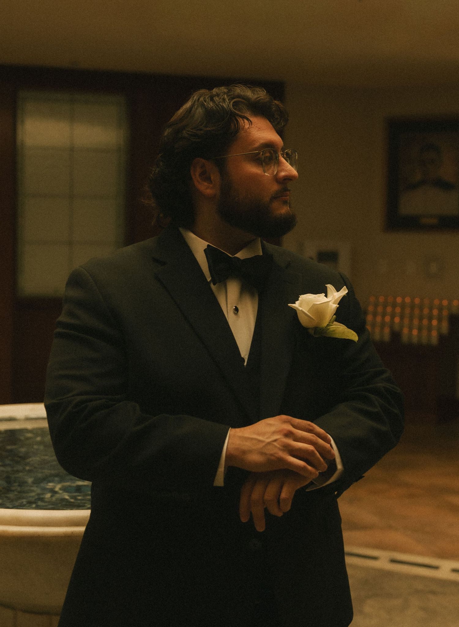 Man in black tuxedo with white boutonniere stands in dimly lit room, hands clasped, looking away.