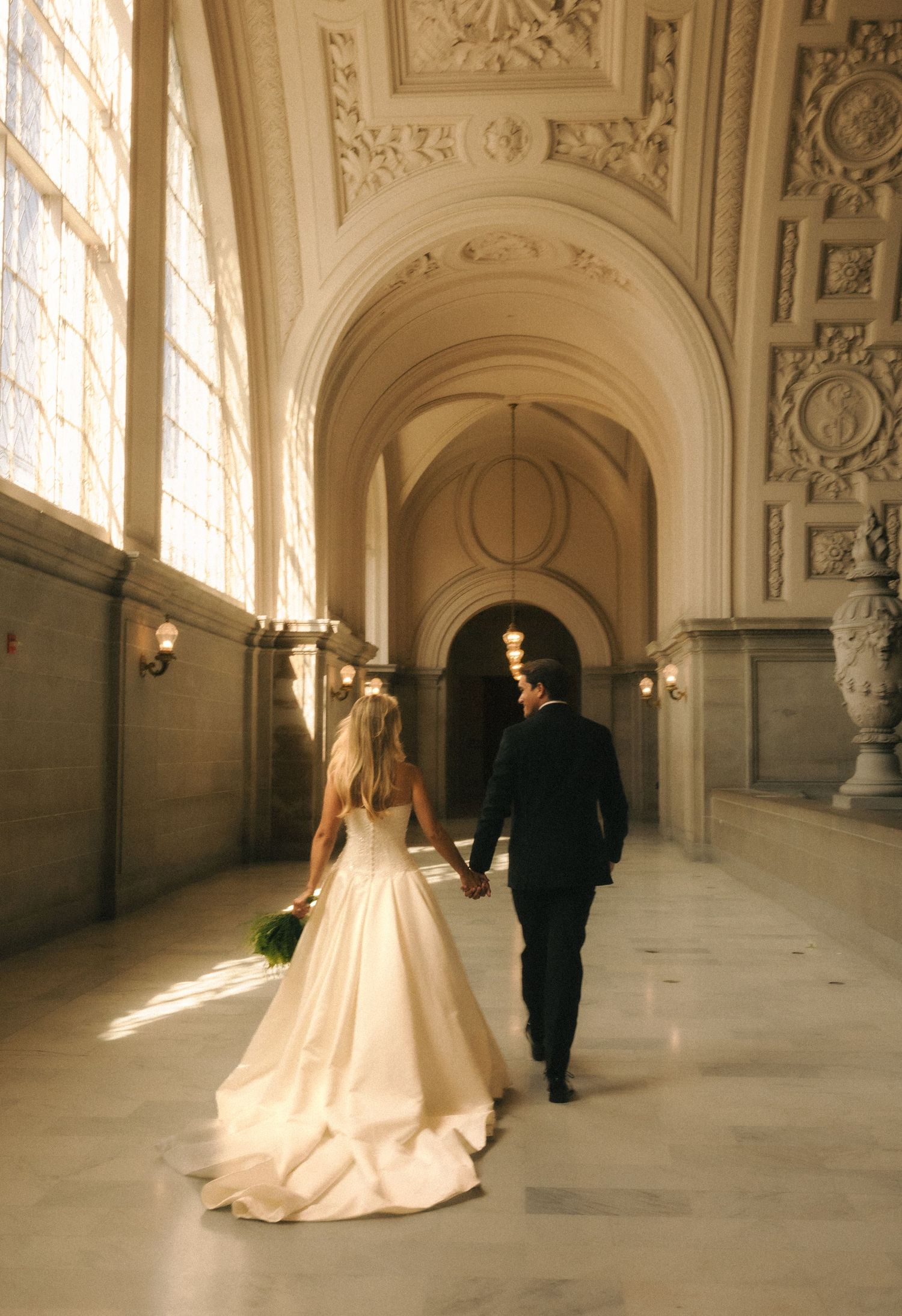 Bride and groom walking hand in hand through an elegant ornate hallway with arched ceilings and golden light.