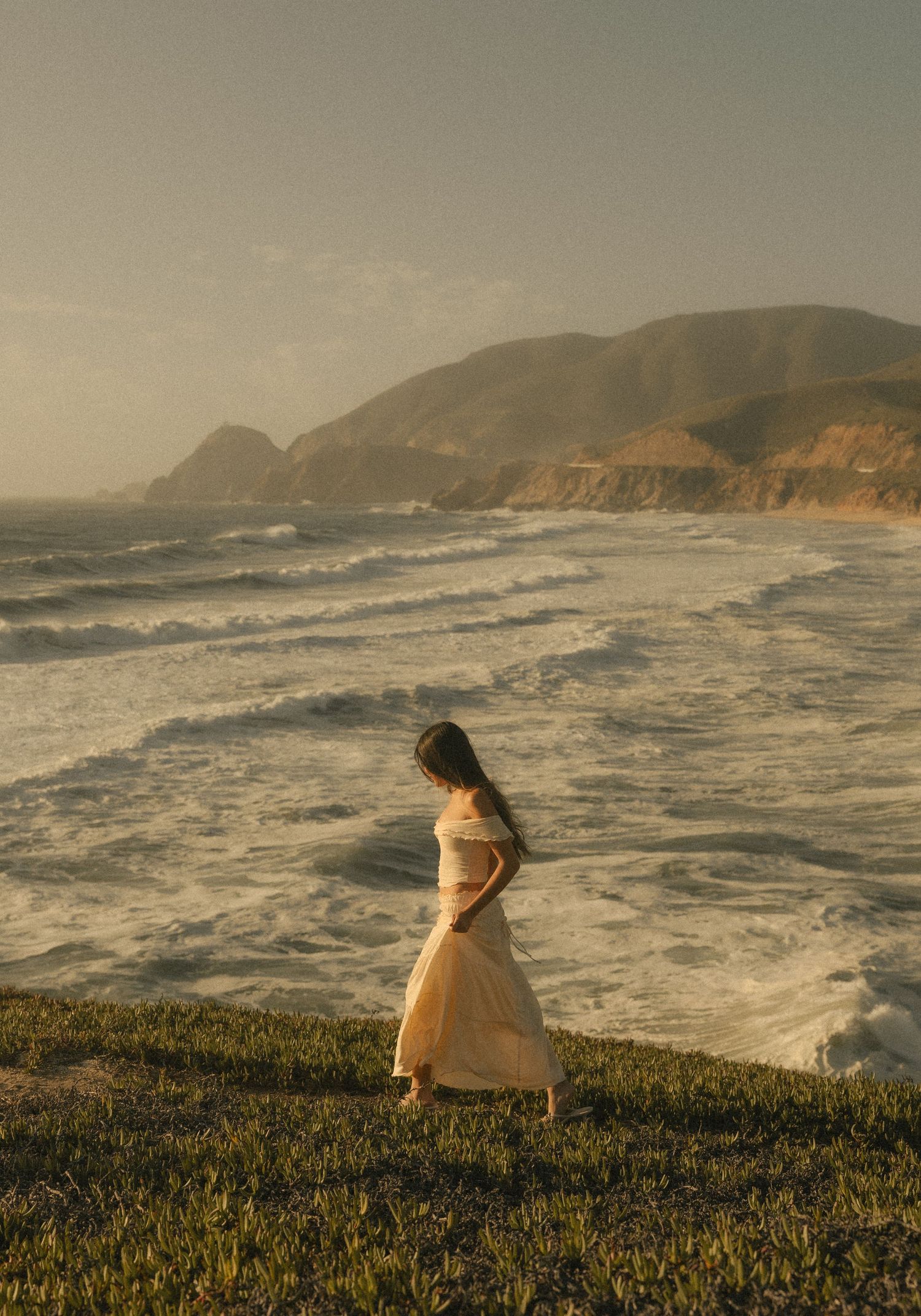 Woman in white dress standing on grassy cliff overlooking dramatic ocean waves with coastal mountains at golden hour.