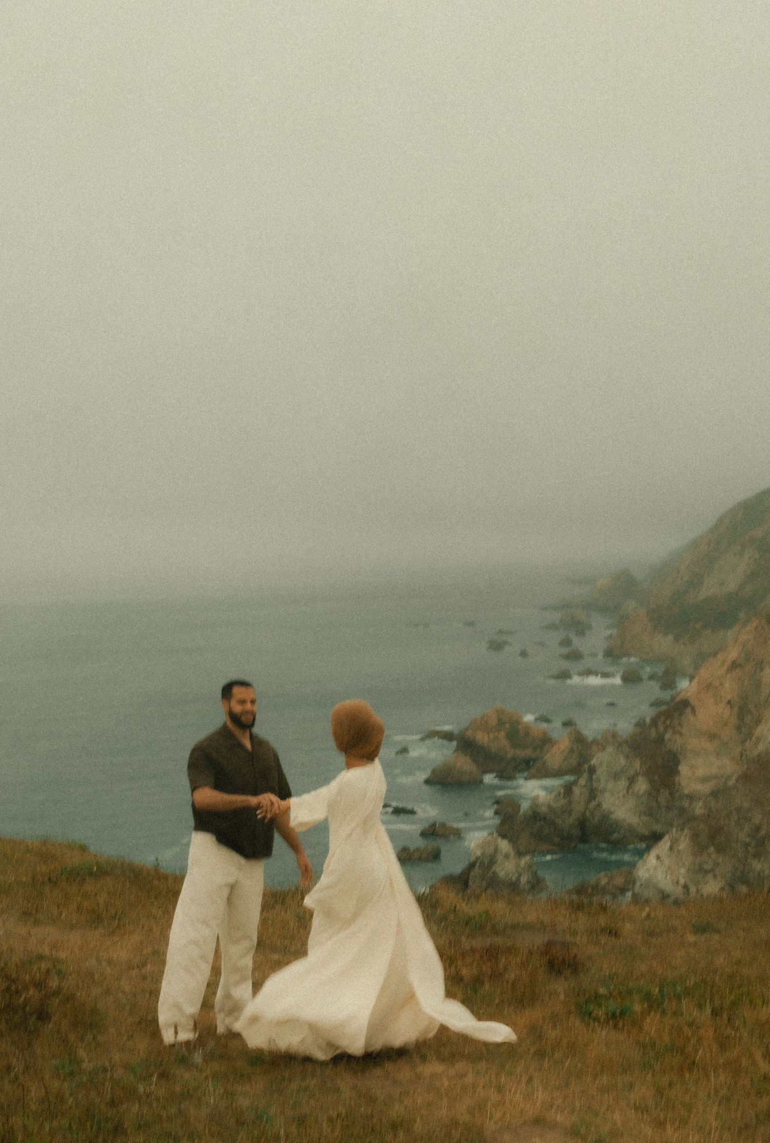 Couple holding hands on a coastal cliff overlooking rocky shores and misty ocean, woman in white dress, man in dark shirt.
