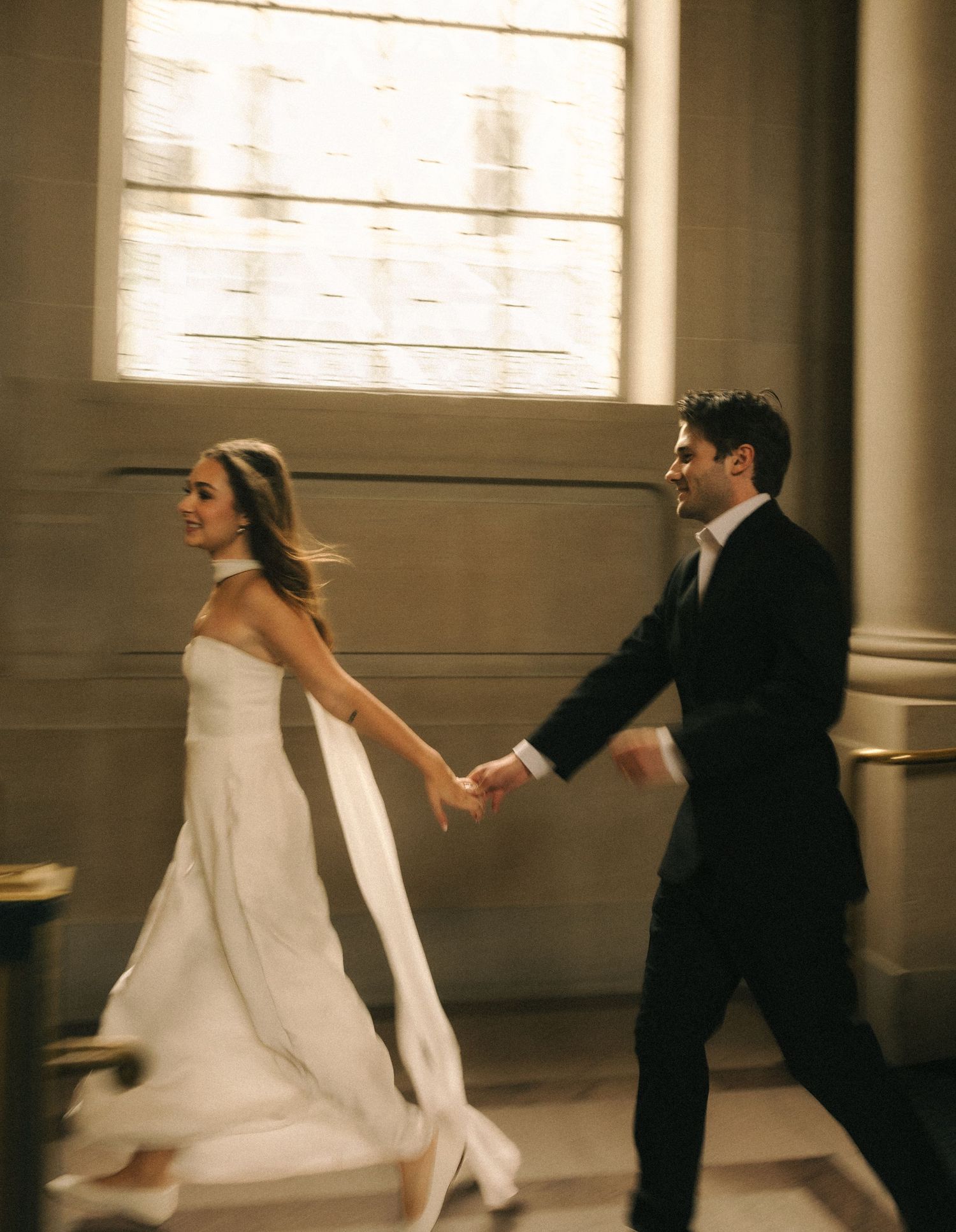A bride in a white gown and groom in a black tuxedo hold hands while walking through a sunlit elegant venue.