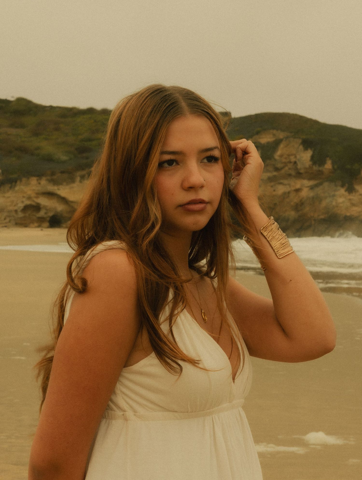 Young woman in white dress poses on sandy beach with ocean waves and green cliffs in background.