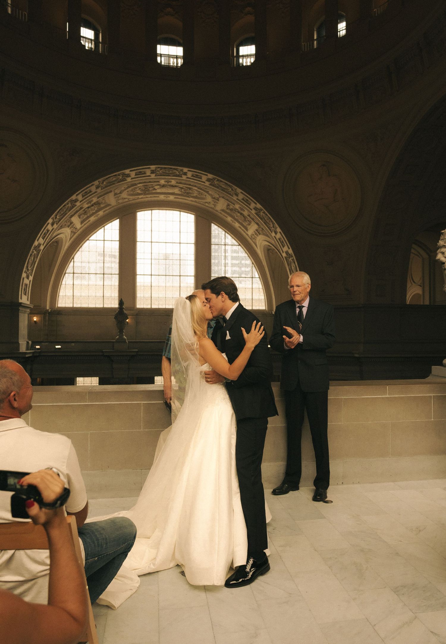 Bride and groom share first kiss at elegant domed wedding venue while photographer captures the romantic ceremony moment.