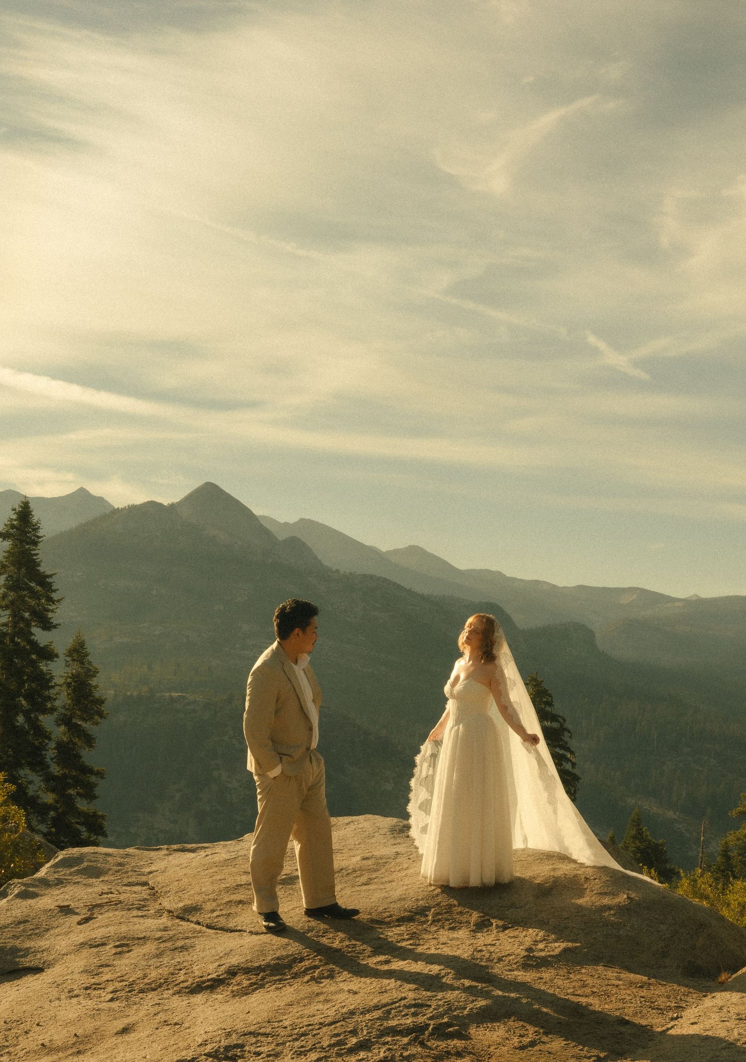 Bride in white gown and groom in tan suit stand on mountain overlook with scenic valley and peaks behind them.
