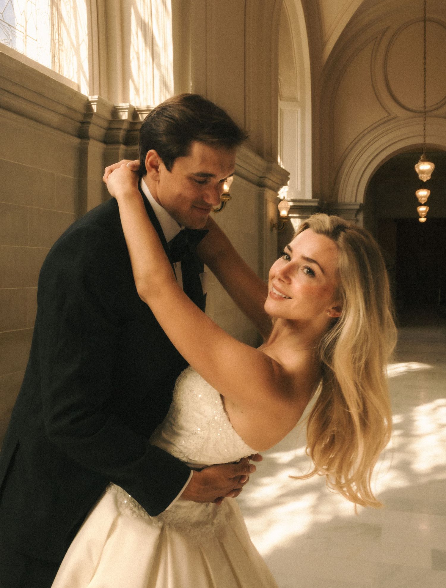 Elegant bride and groom sharing a romantic dance in a grand hall with arched columns and warm golden lighting.