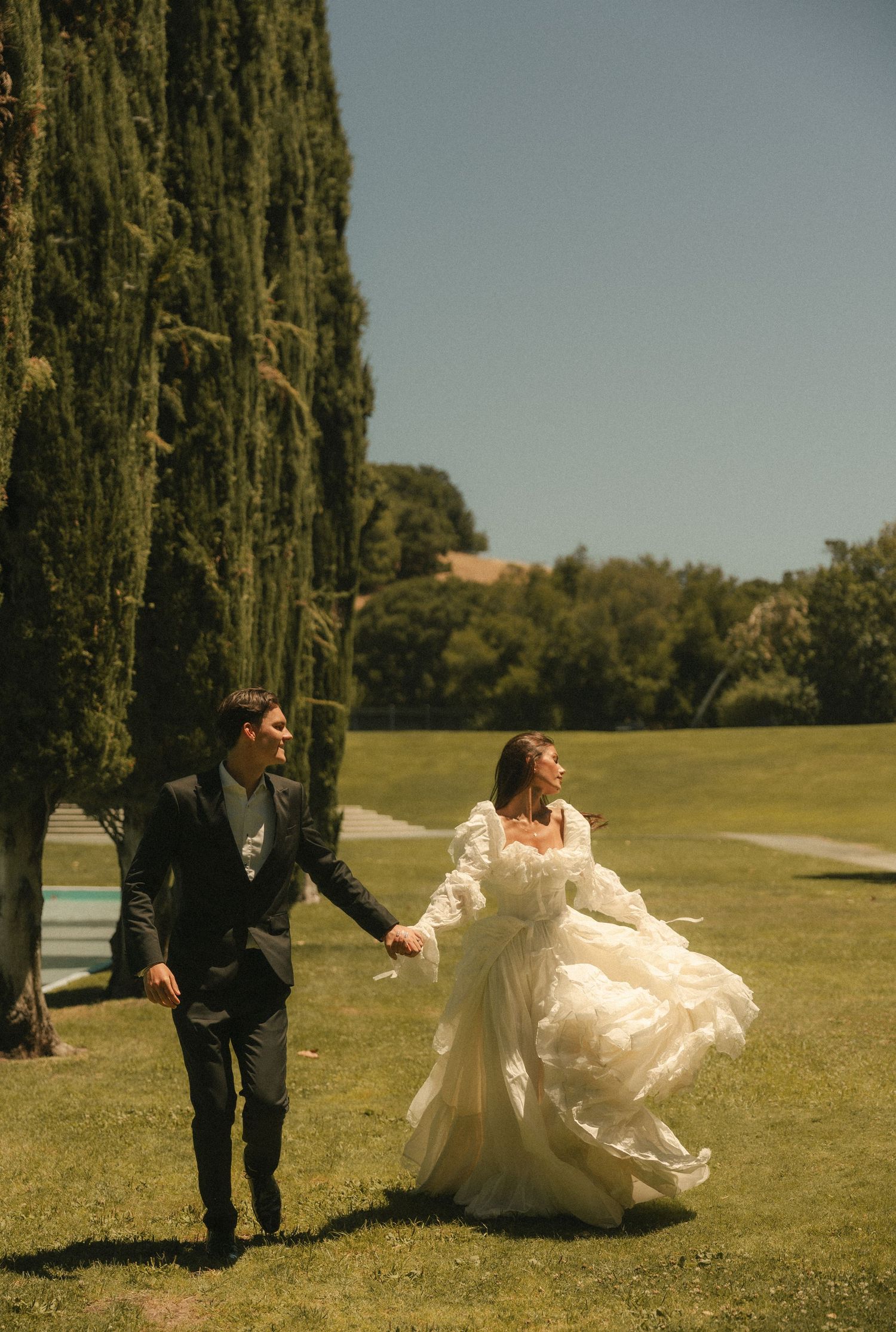Bride in flowing white gown and groom in dark suit hold hands walking across green lawn with tall cypress trees.