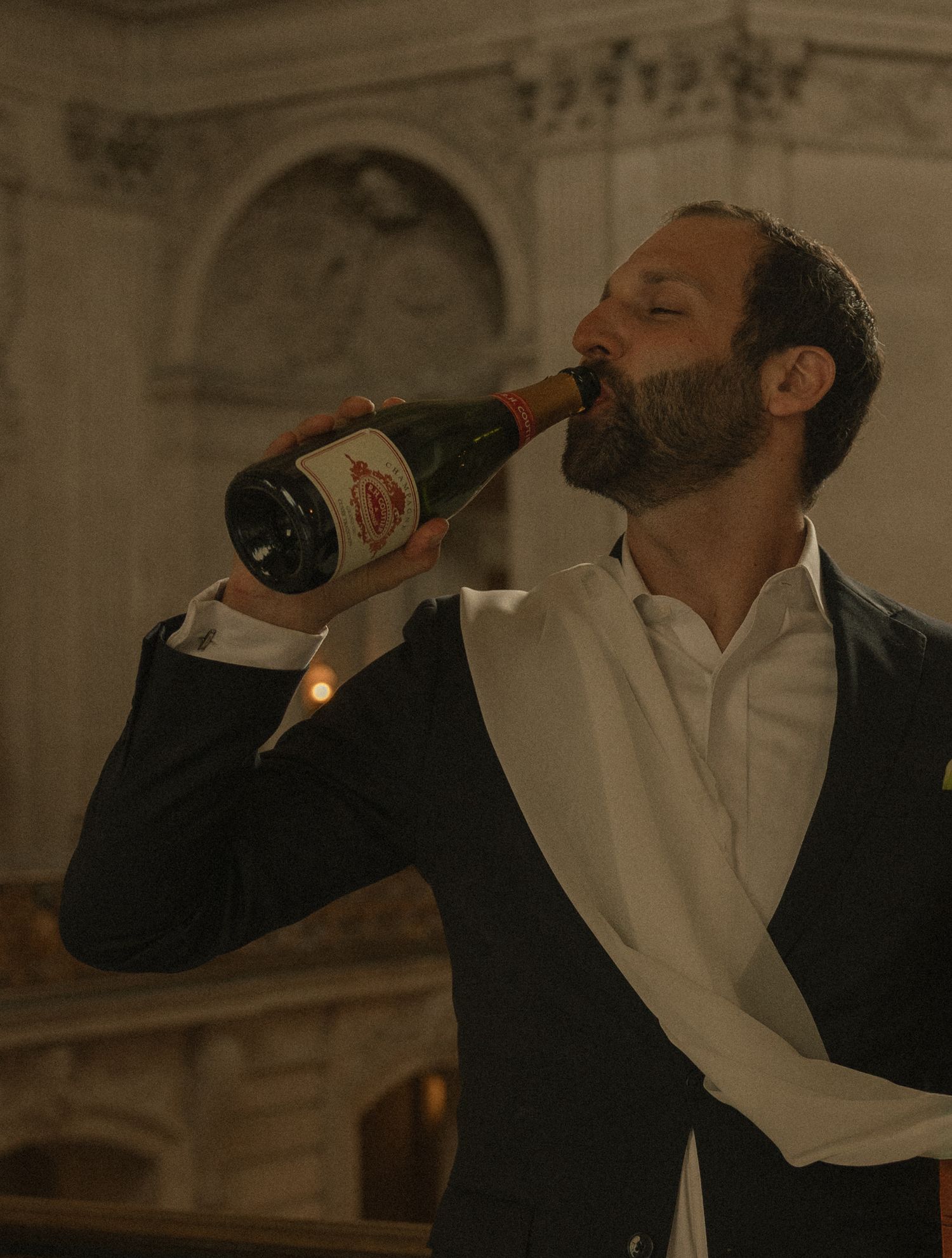 Bearded man in a dark blazer drinking directly from a large wine bottle in an elegant architectural setting.