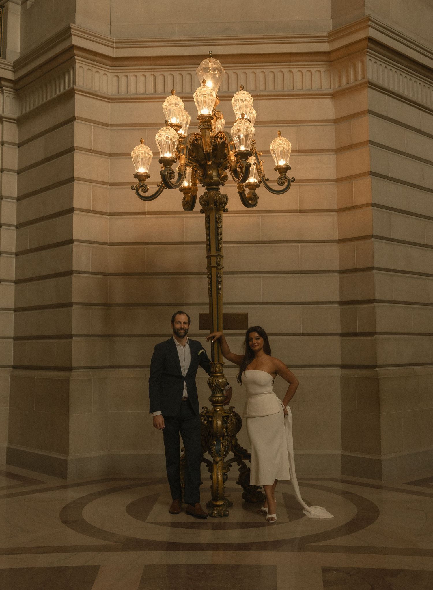 Bride and groom posing under ornate gold candelabra lamp at San Francisco City Hall wedding ceremony.