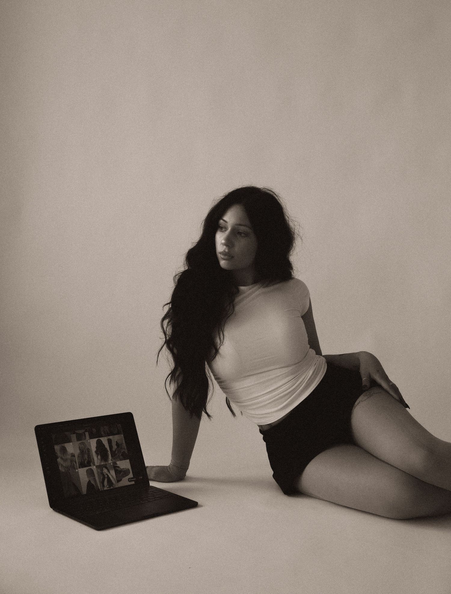 Woman in white top and black shorts sits on floor beside open laptop in monochrome studio photo.